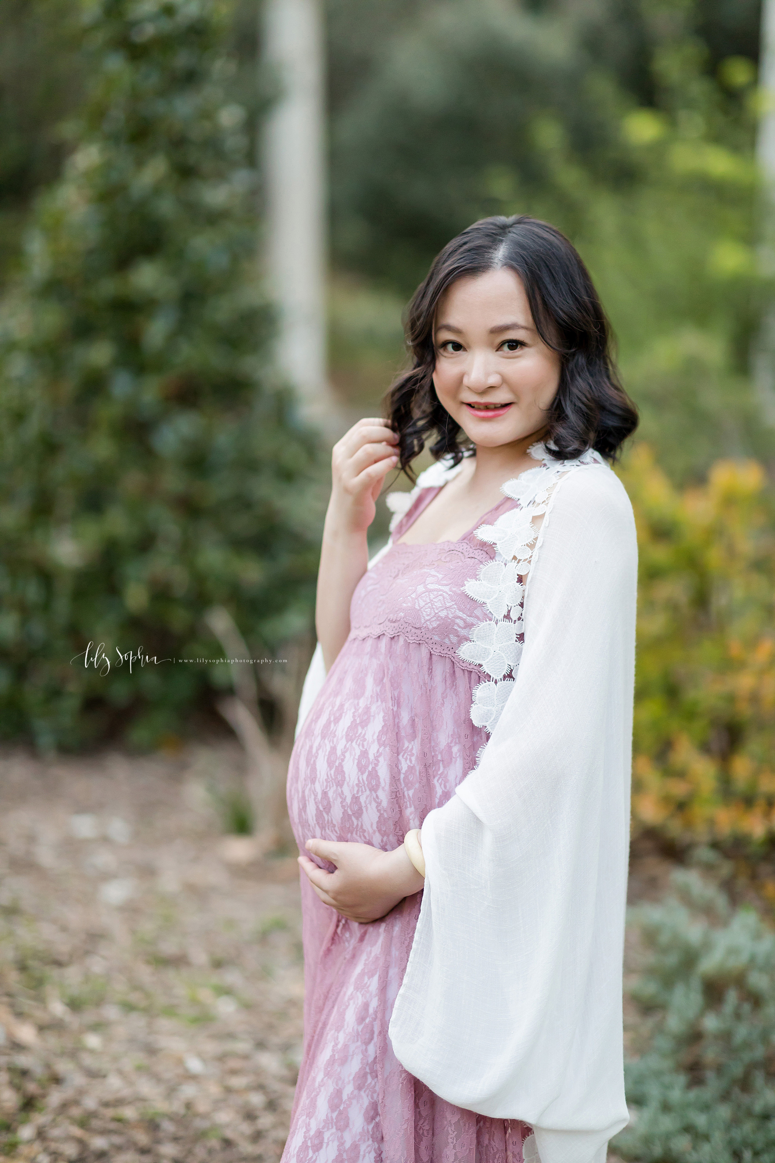  Pregnant woman poses in an Atlanta garden for a photo. She is wearing a rose colored lace full-length sundress and a white shawl with a flowered edge as she stands in the garden. She is playing in her hair with her right hand and holding her belly w