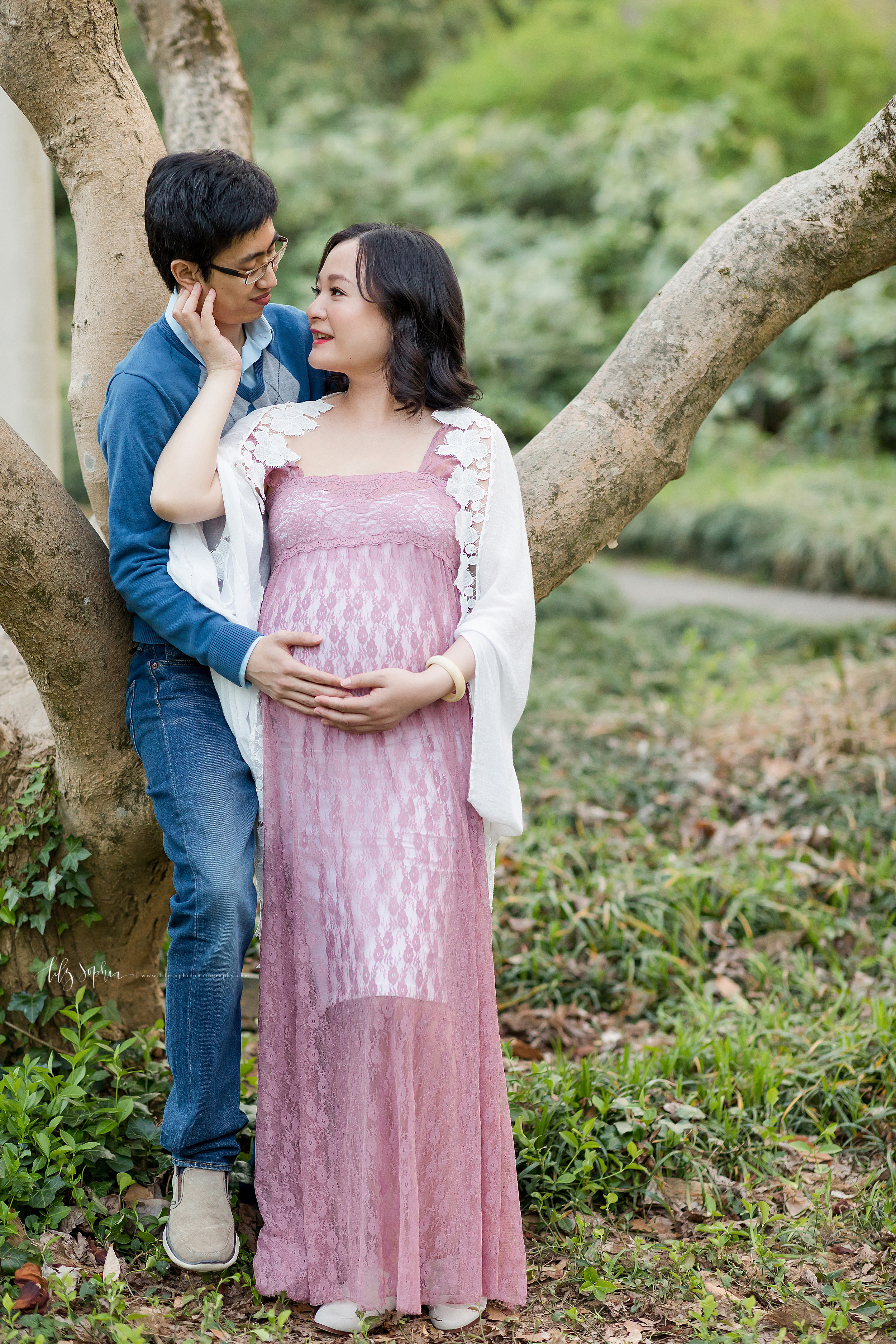  Asian couple’s maternity shoot taken by Lily Sophia Photography in an Atlanta garden. The couple is standing in front of a tree. The husband’s back is against the tree as he turns his head to the right to face his wife. His wife stands in front of h