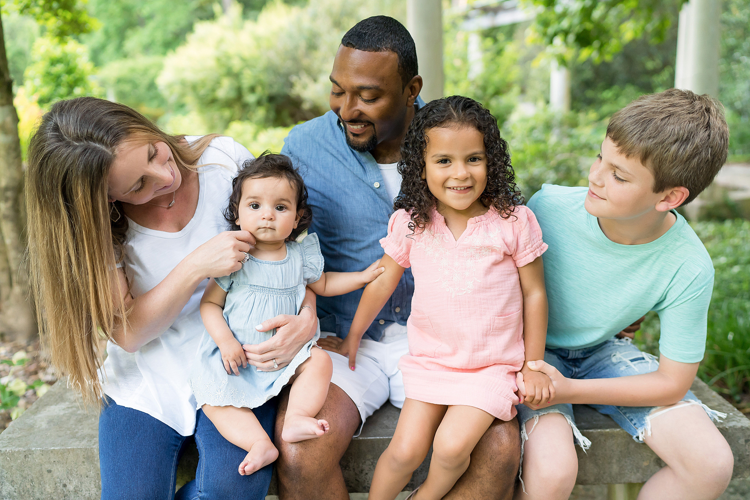  Family photo of five sitting on a stone bench in an Atlanta garden at sunset. Mom is looking at her baby daughter and trying to get her to smile by tickling her near her chin while dad and her brother look on and her older sister smiles. 