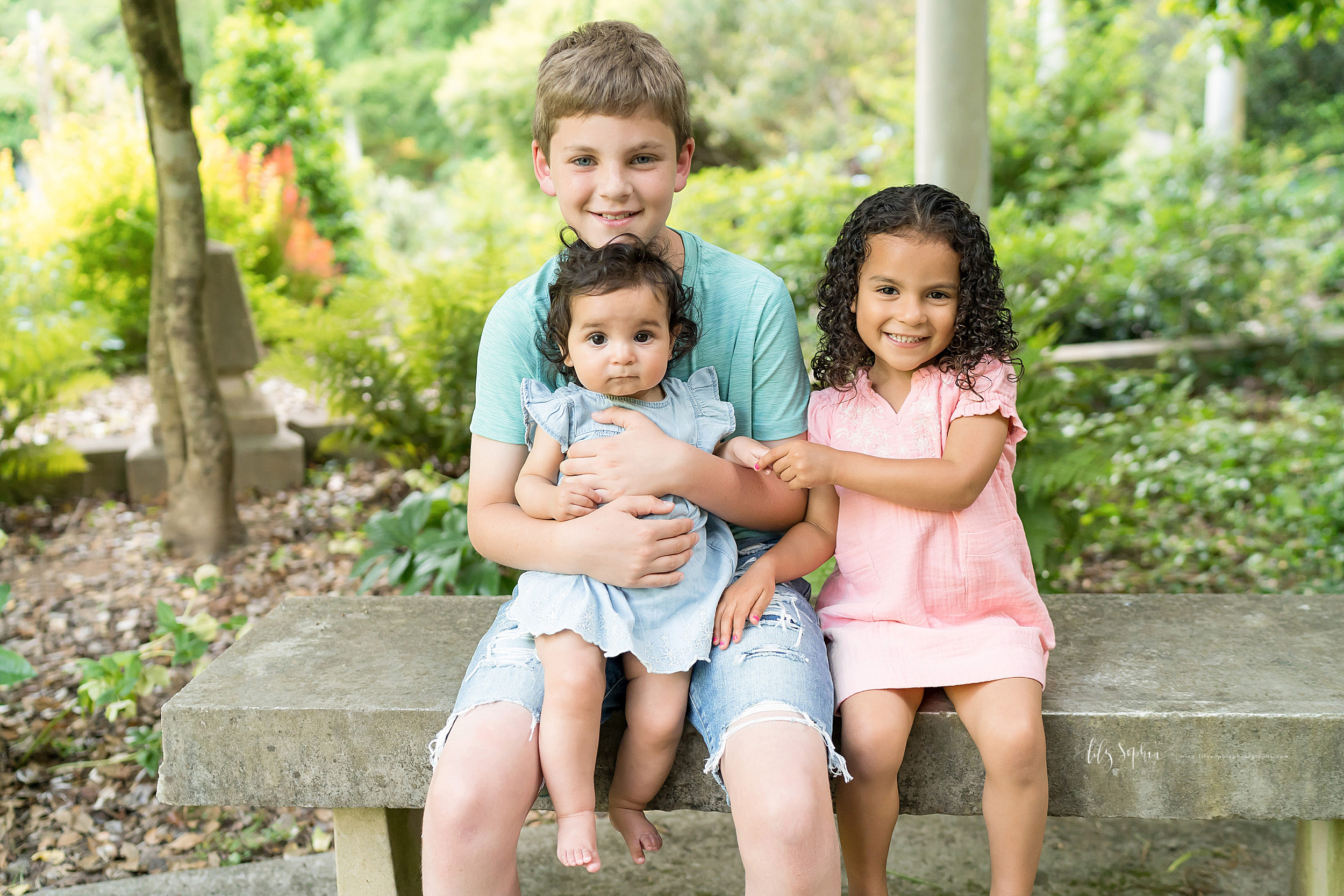  Photo of three happy siblings sitting in an Atlanta garden.  The brother and two sisters are sitting on a cement bench.  The older brother is holding his baby sister on his lap.  His younger sister is sitting next to him and is holding her baby sist