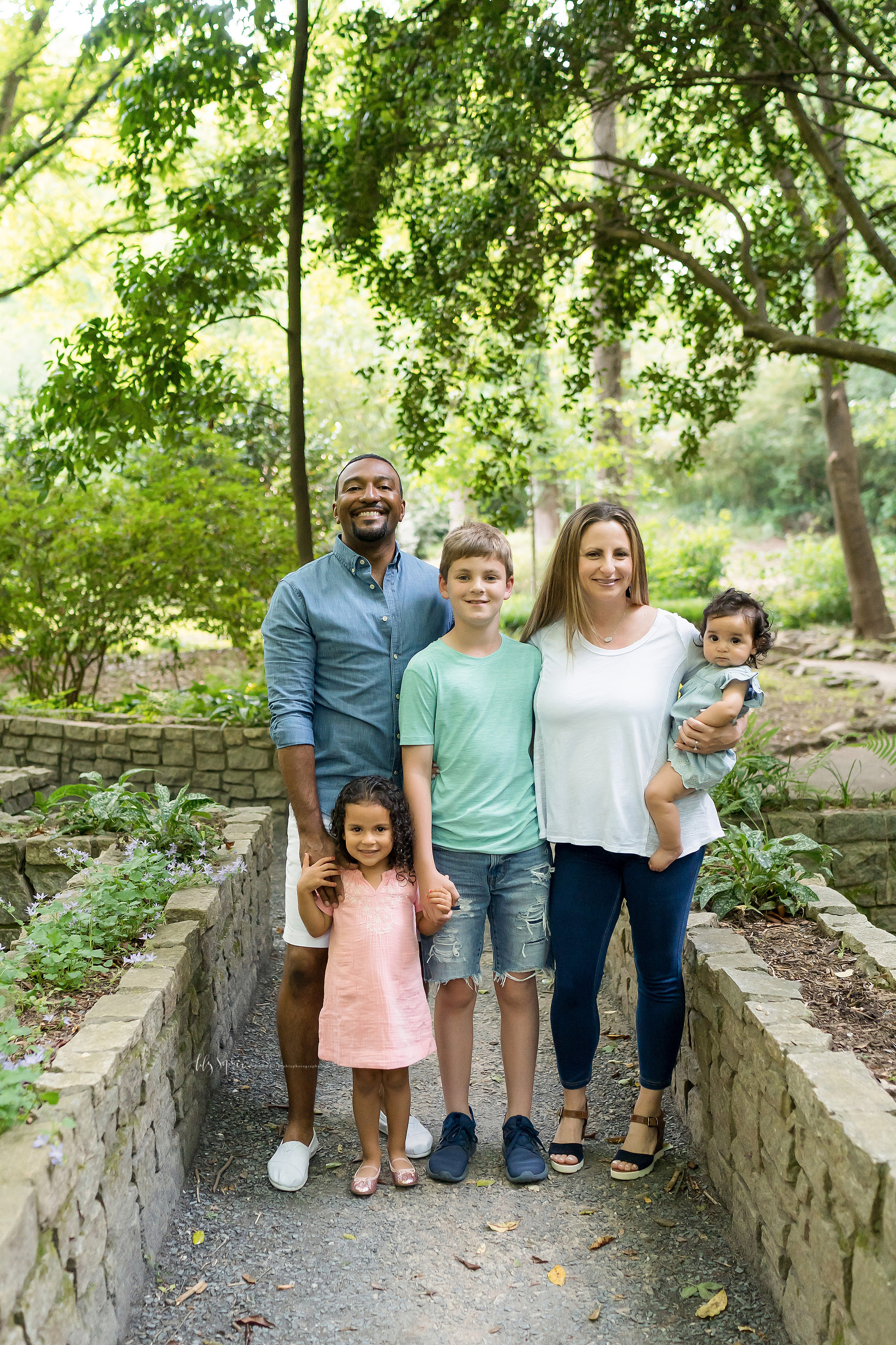  Family photo session in an Atlanta garden with a happy family of five. The mom and son are in the center of the photo as they stand next to one another. Dad is standing behind his son and has his right hand on his young daughter’s right shoulder. Th