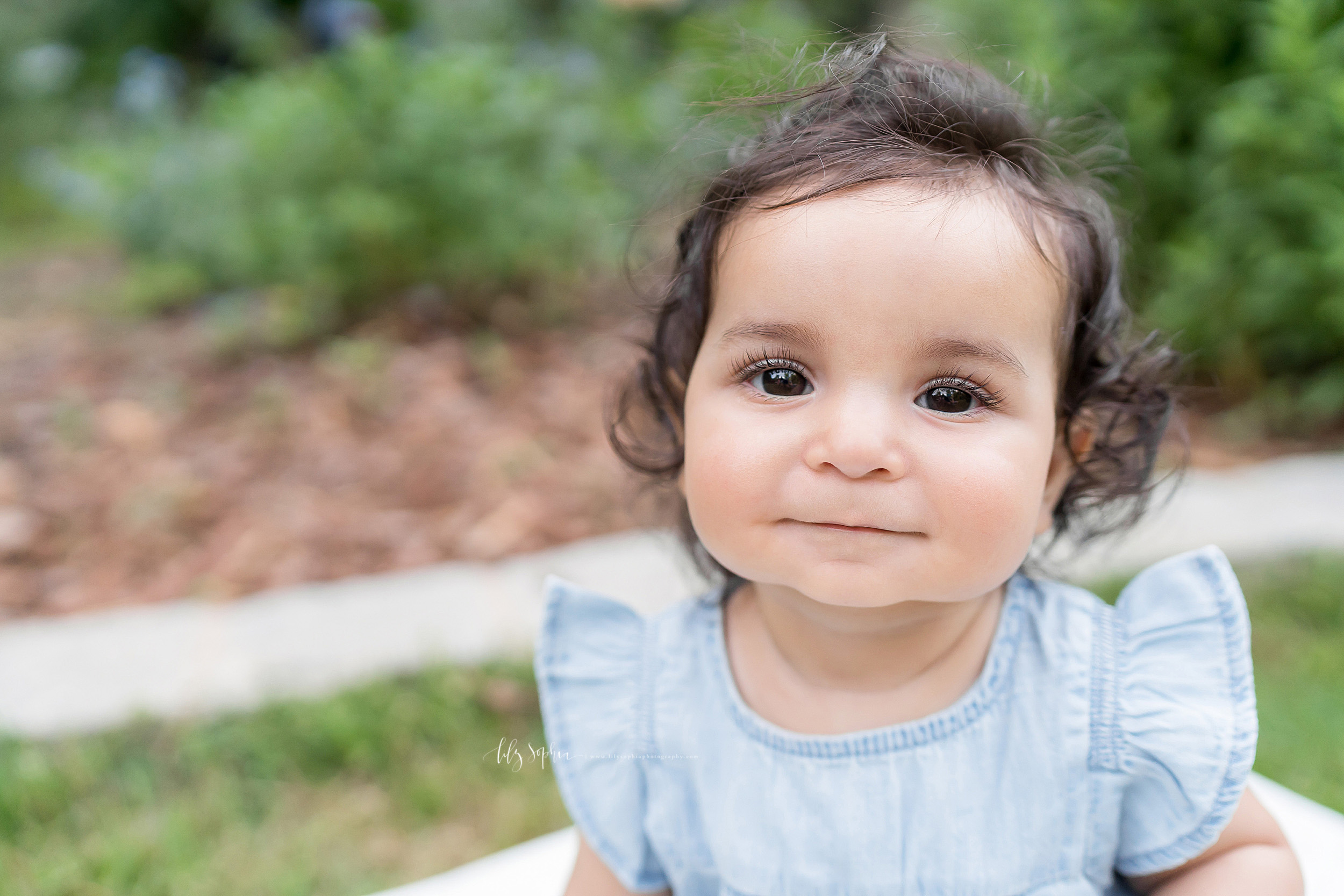  Photo of a precious baby girl as she sits in an Atlanta garden taken by Lily Sophia Photography.  The brown haired, brown eyed baby is grinning.  Her long eyelashes highlight her bright brown eyes.  She is wearing a caped sleeved light blue scooped 