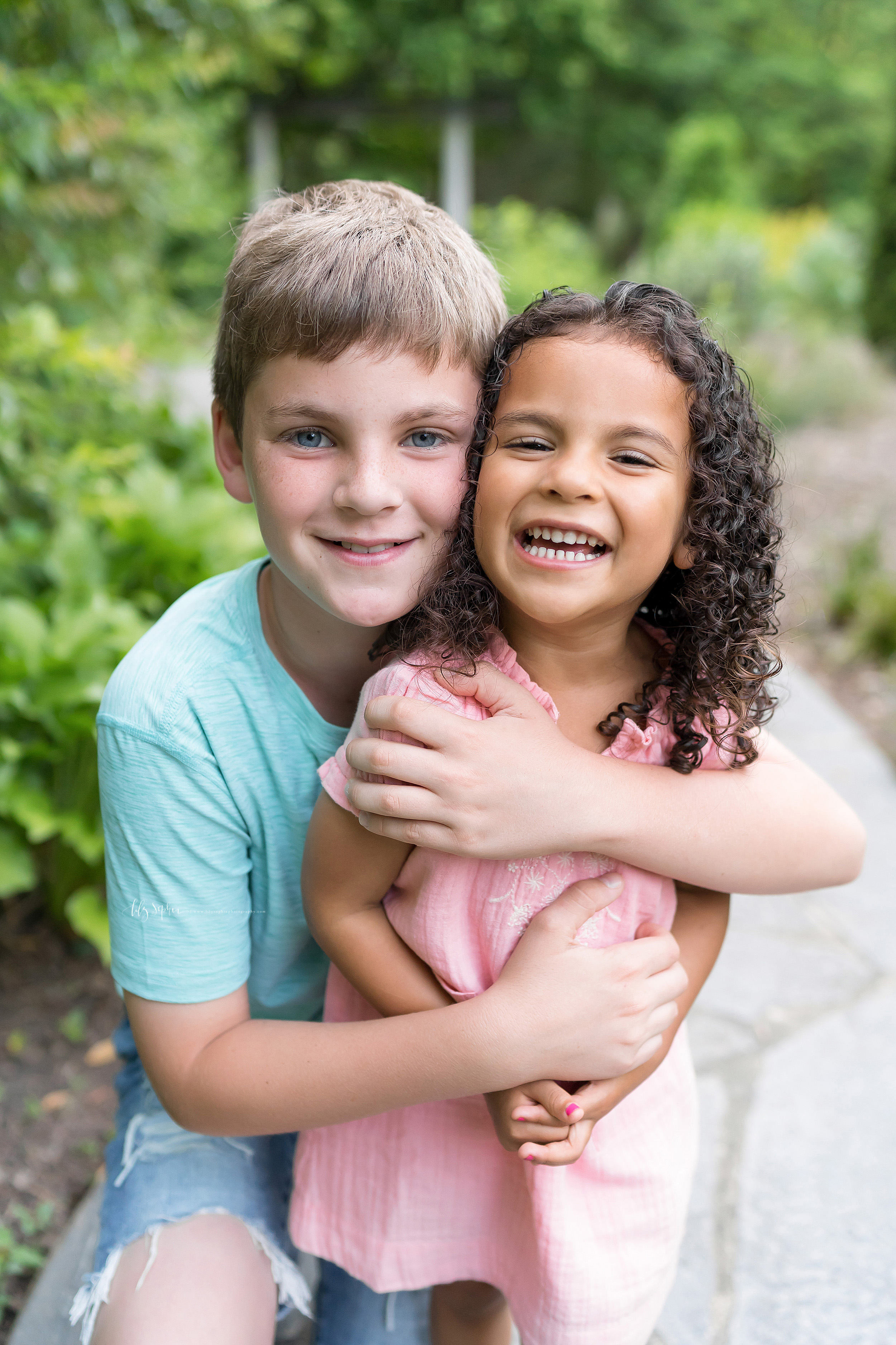  Photo of happy siblings in an Atlanta garden.  The blond haired, blue eyed, fair skinned brother has his hands wrapped around his brown, curly haired sister as he smiles and she giggles.   