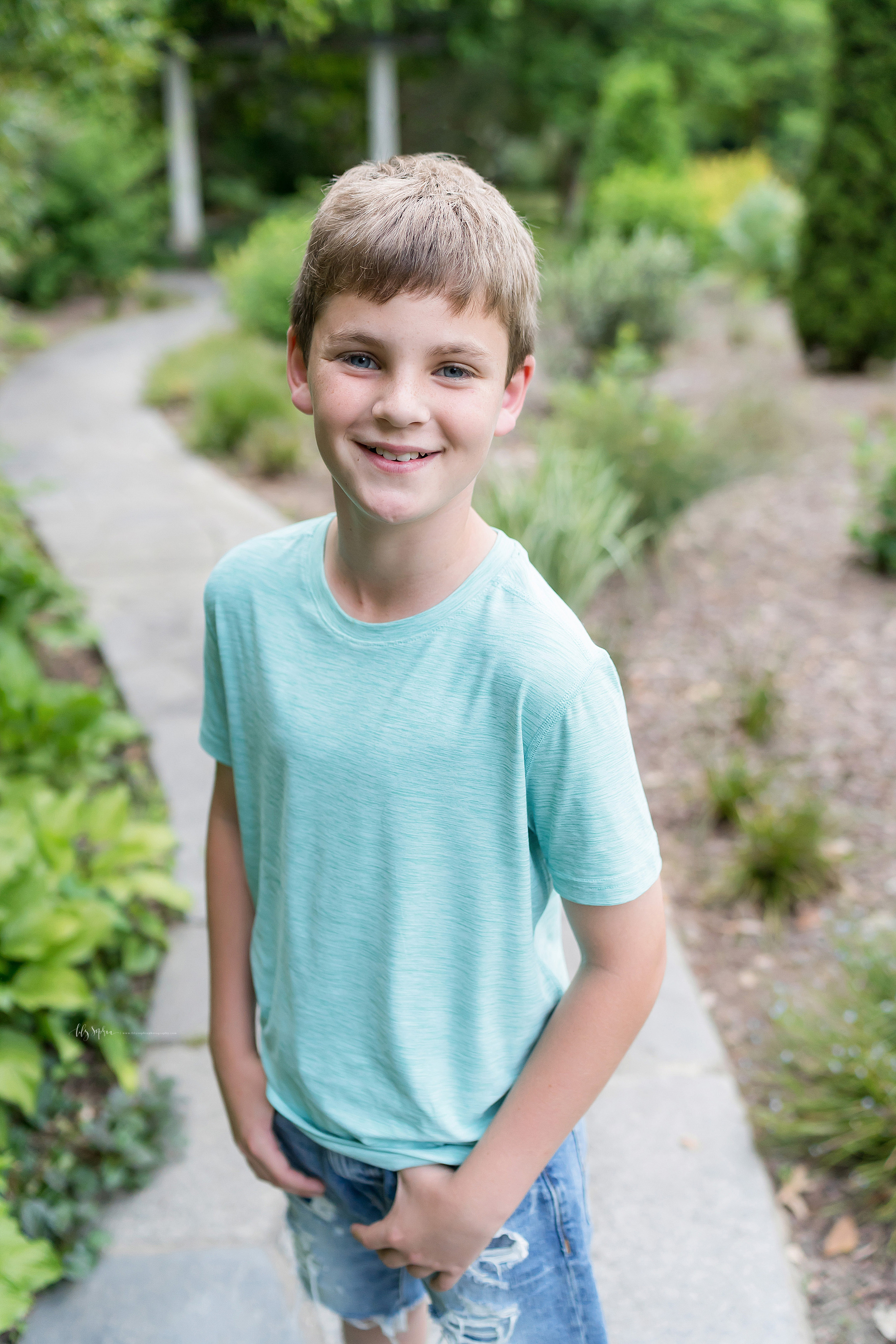  Photo of a smiling boy in a garden in Atlanta.  The blond haired, blue eyed boy is standing on a concrete path.  He is wearing a mint green T-shirt and jean shorts.  He has his thumbs in his front pockets and he is smiling. 