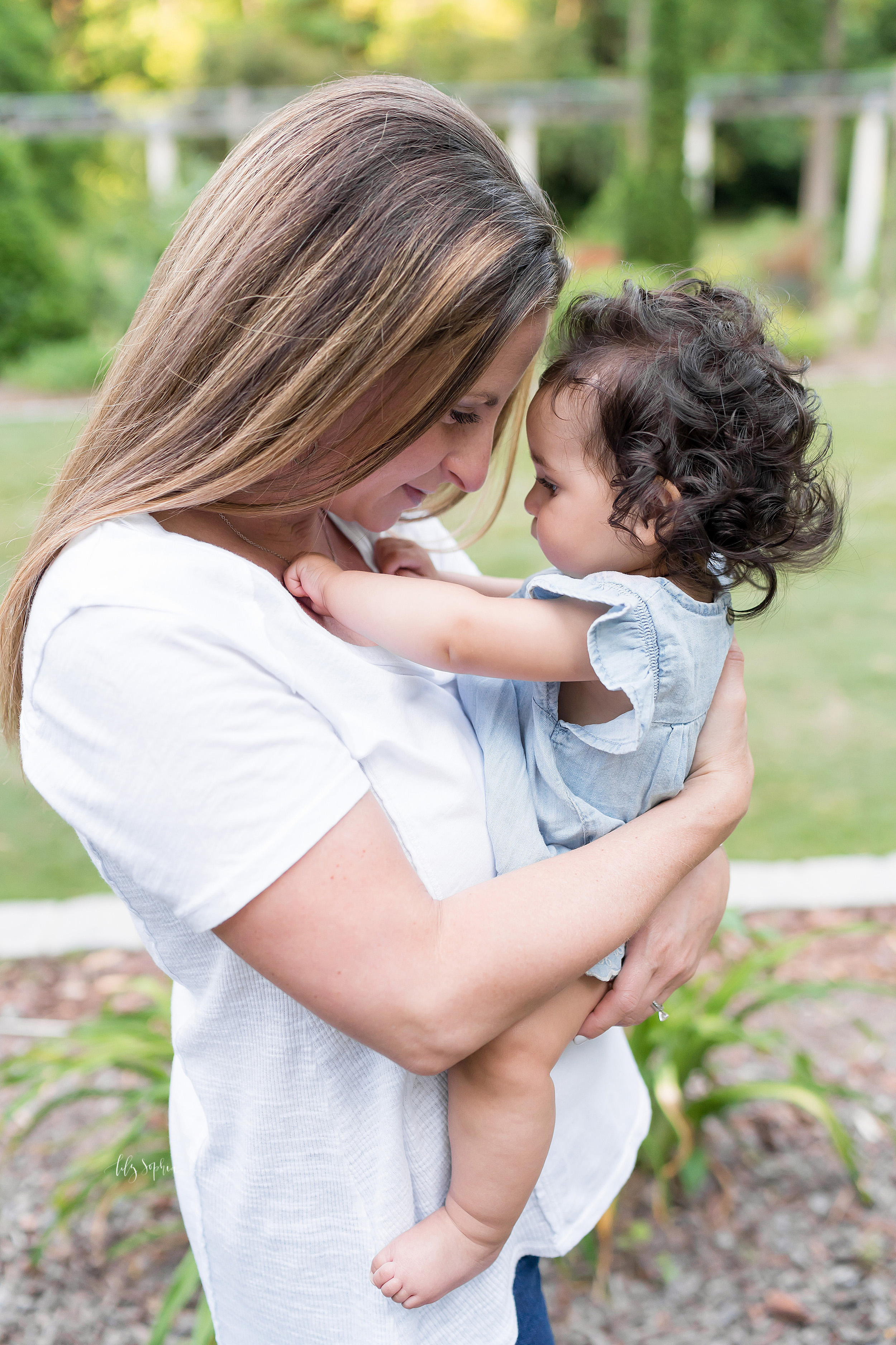  Photo of a mom and her daughter in an Atlanta garden. Mom is holding her barefoot baby girl as she plays with her mom’s necklace. The long blond haired mom is looking down at her daughter. Mom is wearing a short sleeve white V neck T-shirt and jeans