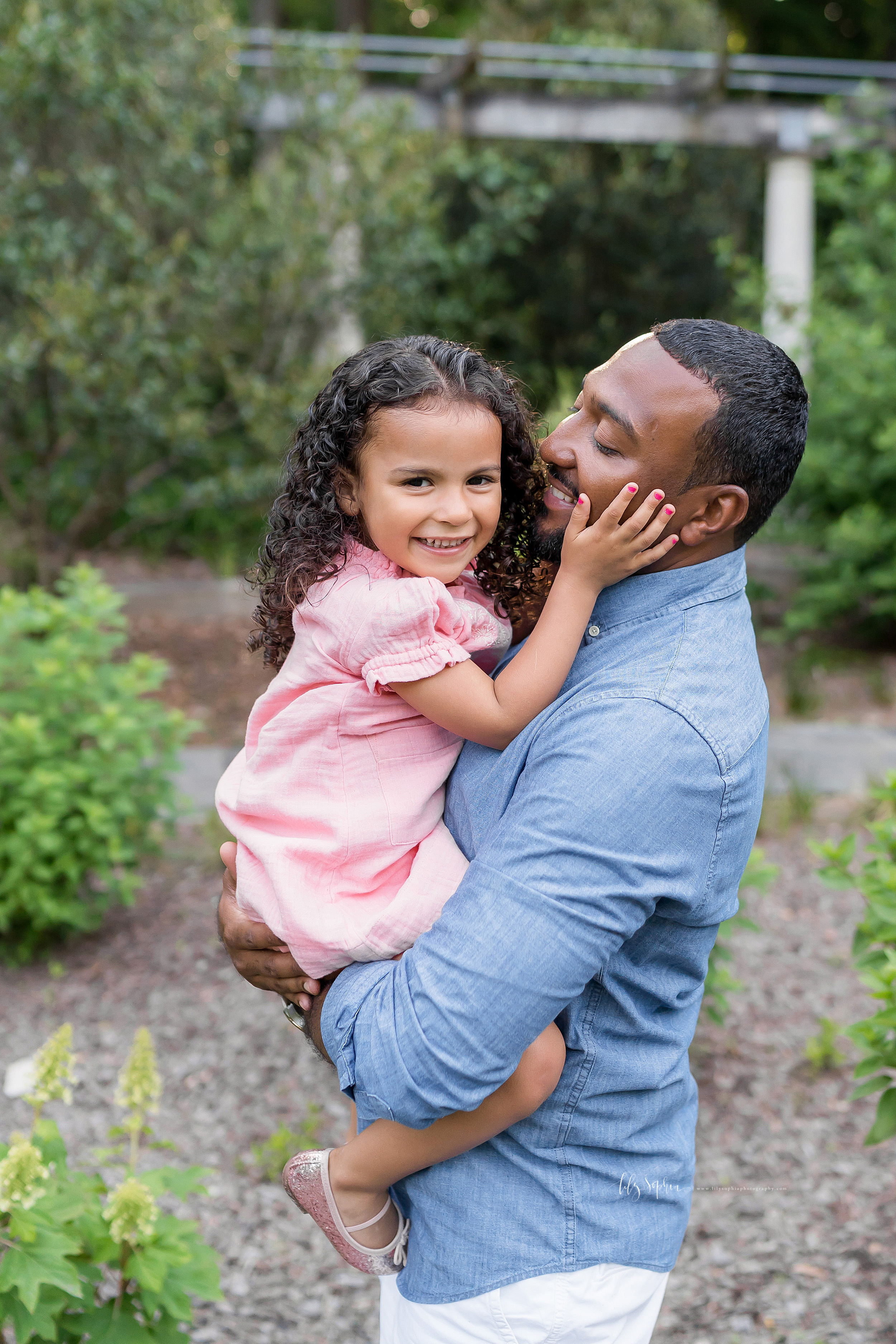  Photo of a happy African-American father holding his young daughter in an Atlanta garden. The father is showcasing his profile as he holds his daughter in his arms. The young daughter has her hands on her father’s cheeks and you can see her delicate