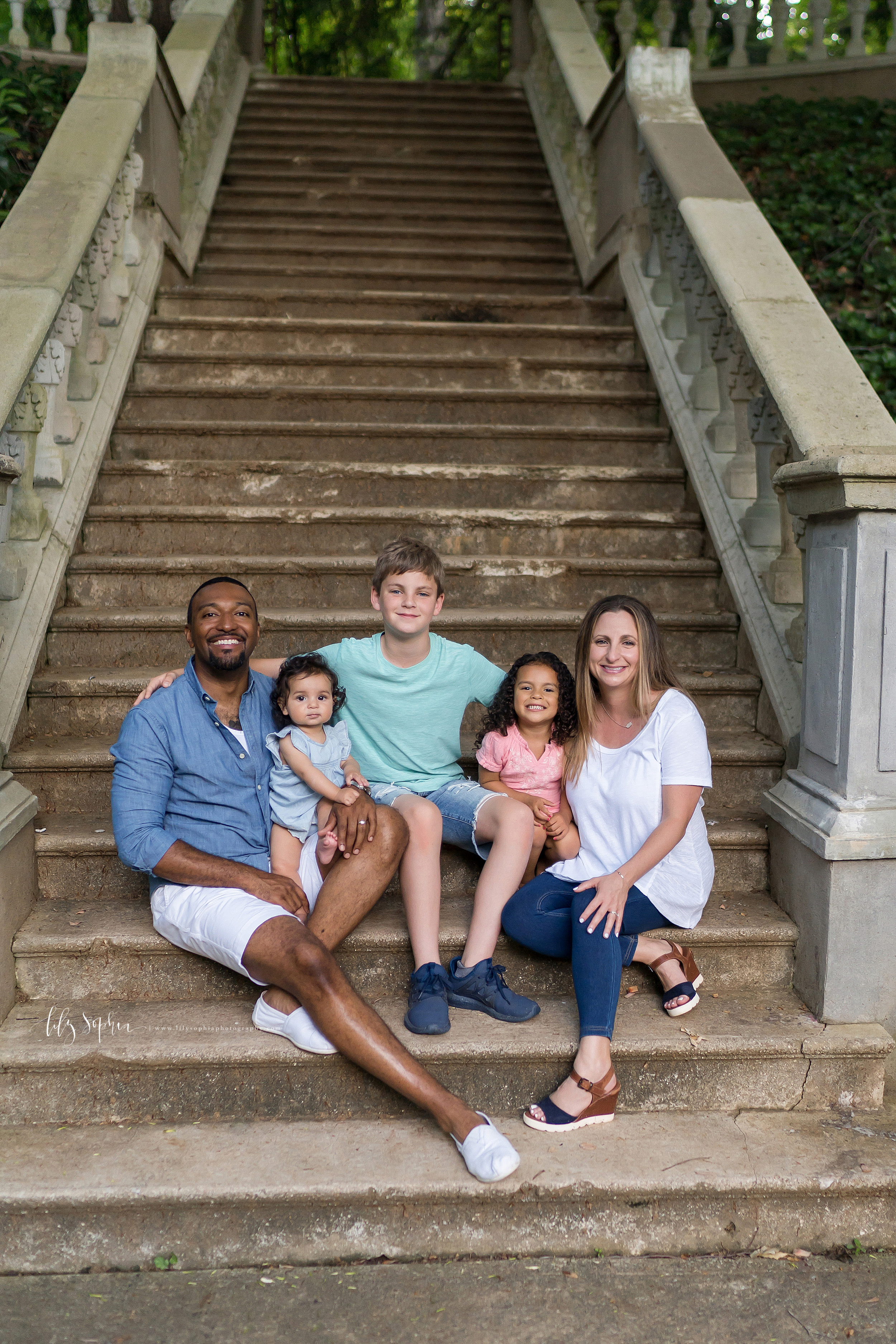  Photo of  a happy family of five in an Atlanta garden.  The family of five sits near the bottom of a staircase.  The African-American dad is on the left with his baby girl on his lap.  His tween son is sitting in the center of the photo with his lit