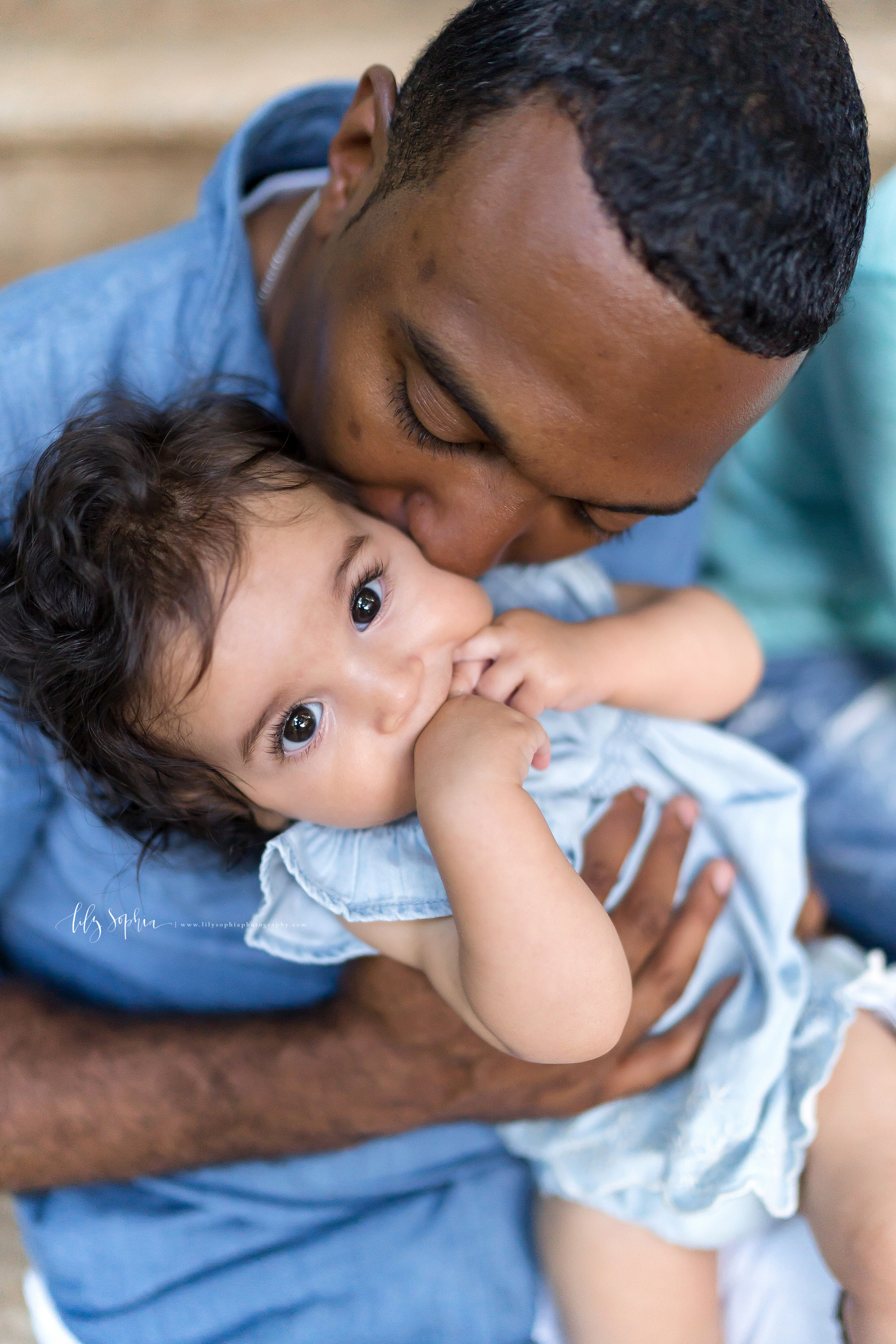  Photo of an African-American father with his baby girl in an Atlanta garden. The father is kissing the cheek of his daughter as he holds her while standing up.  The baby daughter has her fingers in her mouth. 