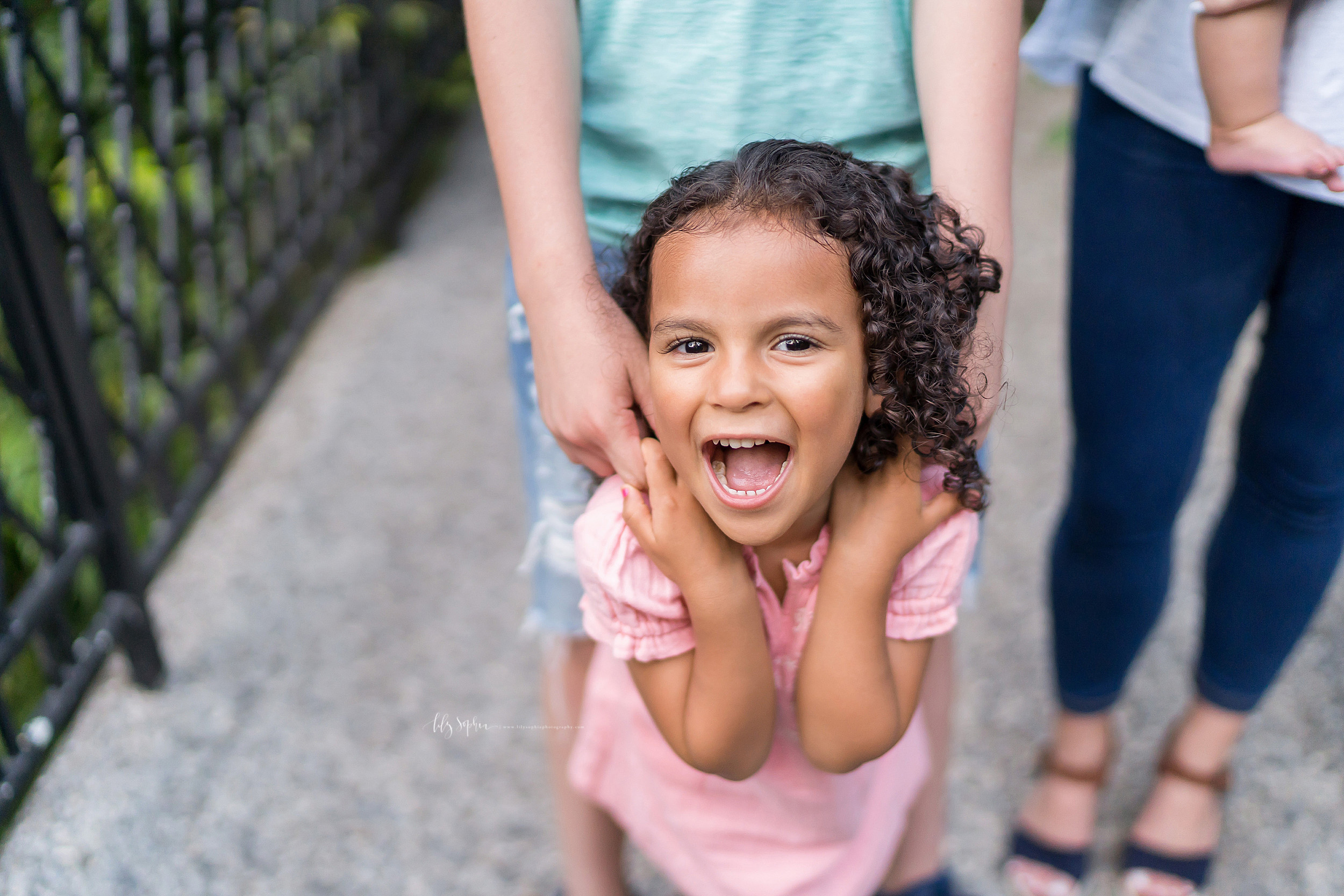  Photo of a laughing young girl in an Atlanta garden. The little girl has her hands up by her shoulders. She is holding her older brother’s fingers as they stand on a bridge. The little girl has her mouth open.  Her mother is standing next to her bro