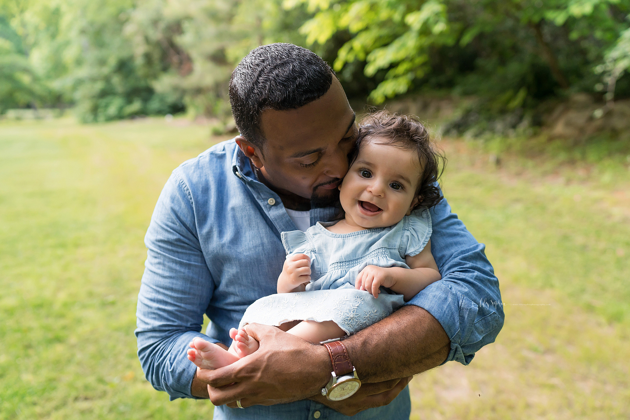  Photo of an African-American dad holding his precious baby girl taken by Lily Sophia Photography in an Atlanta garden.  Dad has his baby girl sitting in his arms as he holds her little feet.  He is whispering in her ear and the little girl is smilin