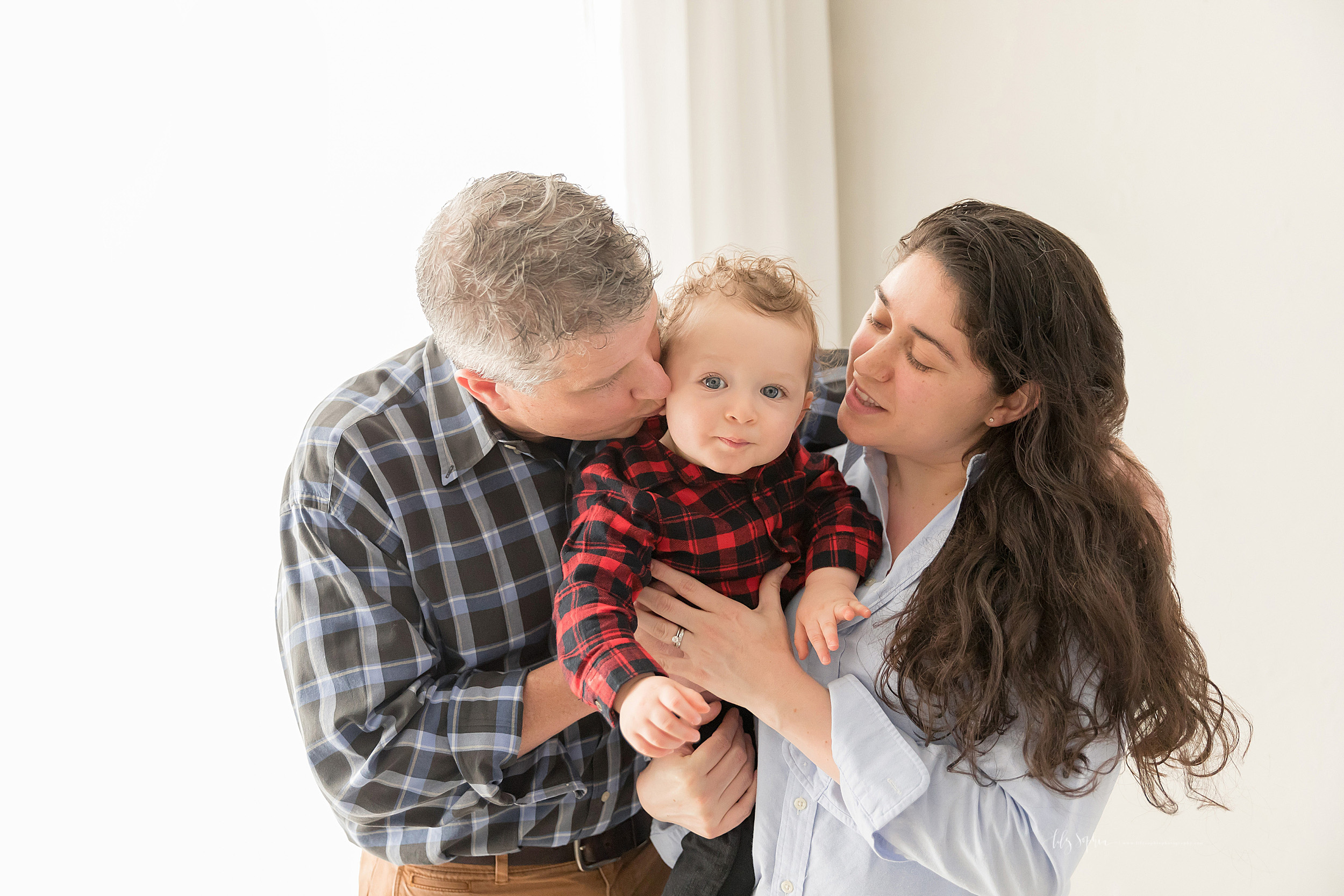  Family photo of a curly haired, blue-eyed one year old with his parents on his first birthday. The photo taken by Lily Sophia Photography captures the love these parents have for their son.  The long-haired brunette mom is holding her son in her arm