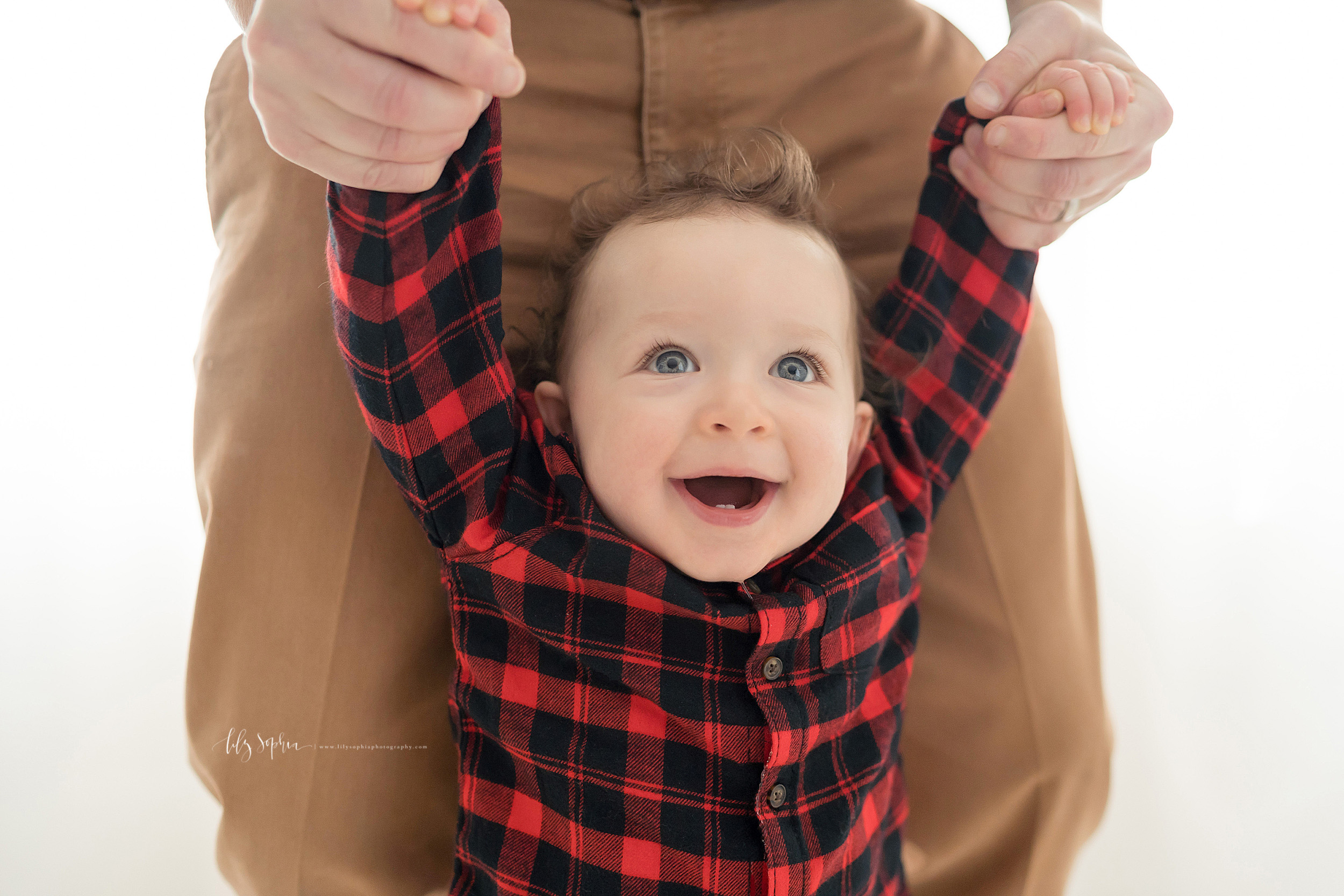 First birthday photo of a one year old as he walks in front of his father.  The curly haired, blue eyed little boy with eyes wide and bright holds his fathers hands to help him walk.  The boy has a smile on his face and you can see his two front tee