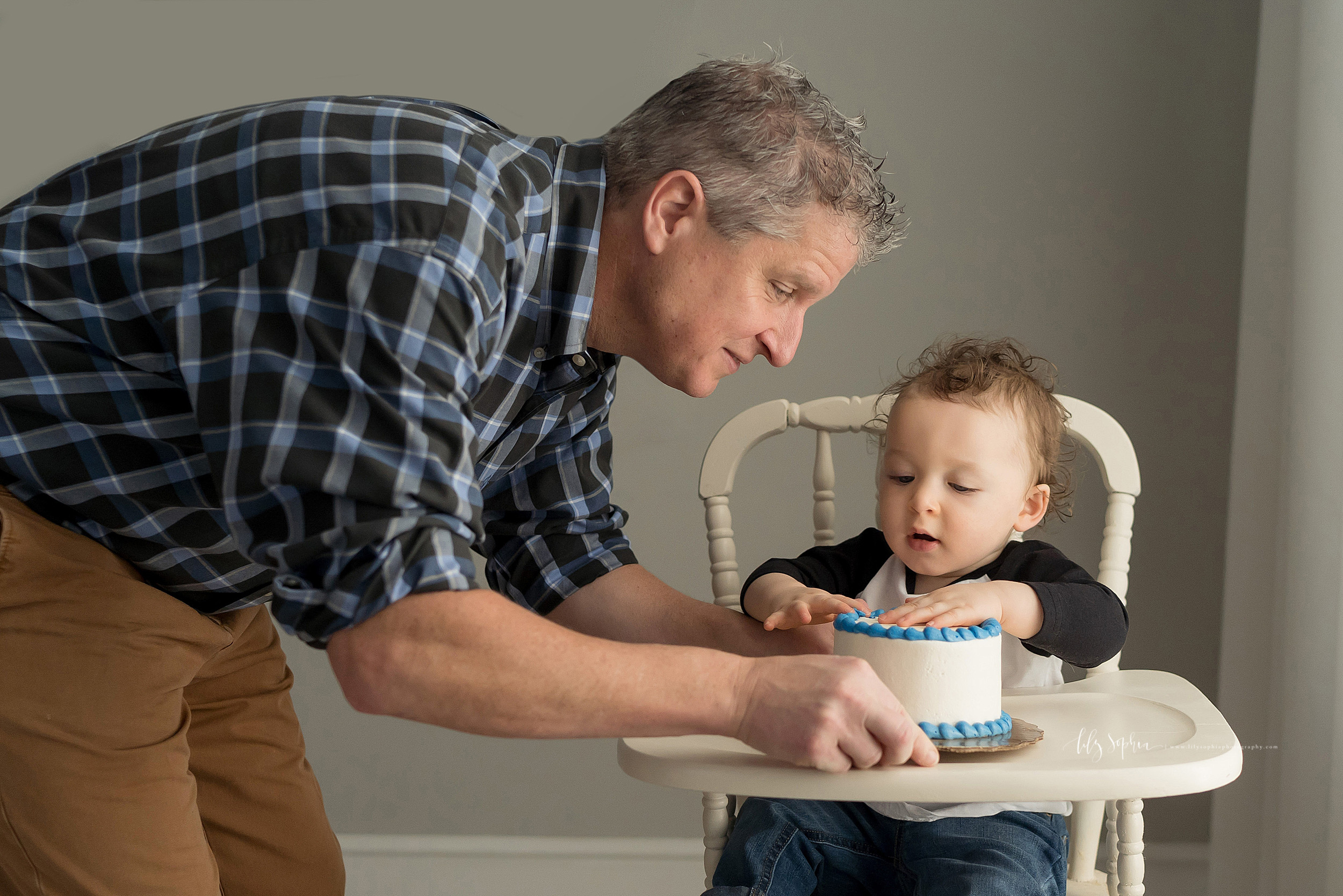  Photo of a father and son on his son’s first birthday. The father is putting a cake in front of his son as his son sits in an antique high chair, pats the cake with his hand, and looks on with anticipation. 