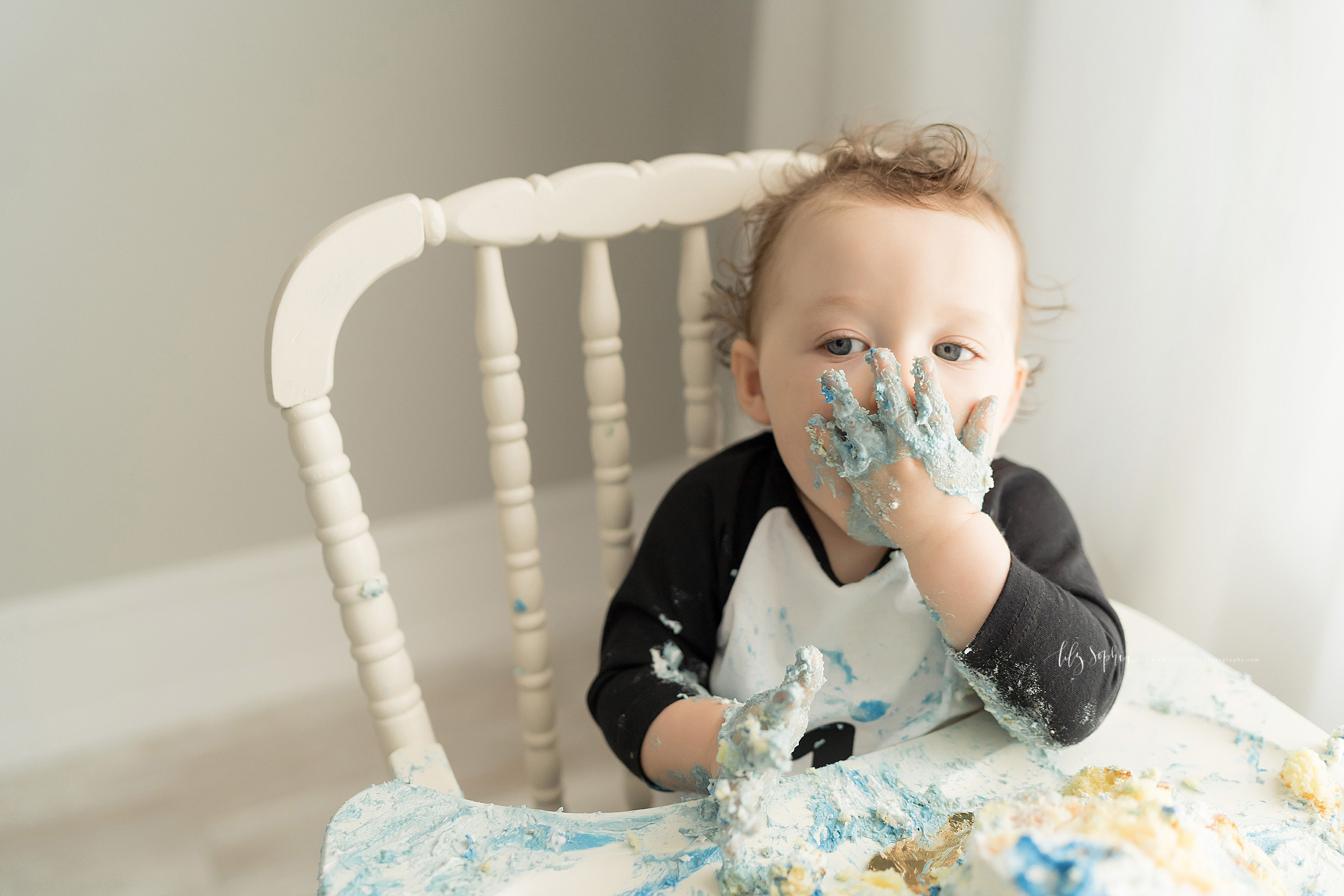  Milestone photo of a baby boy shoving icing and cake into his mouth as he sits in an antique high chair and celebrates turning one in an Atlanta studio in natural light. 