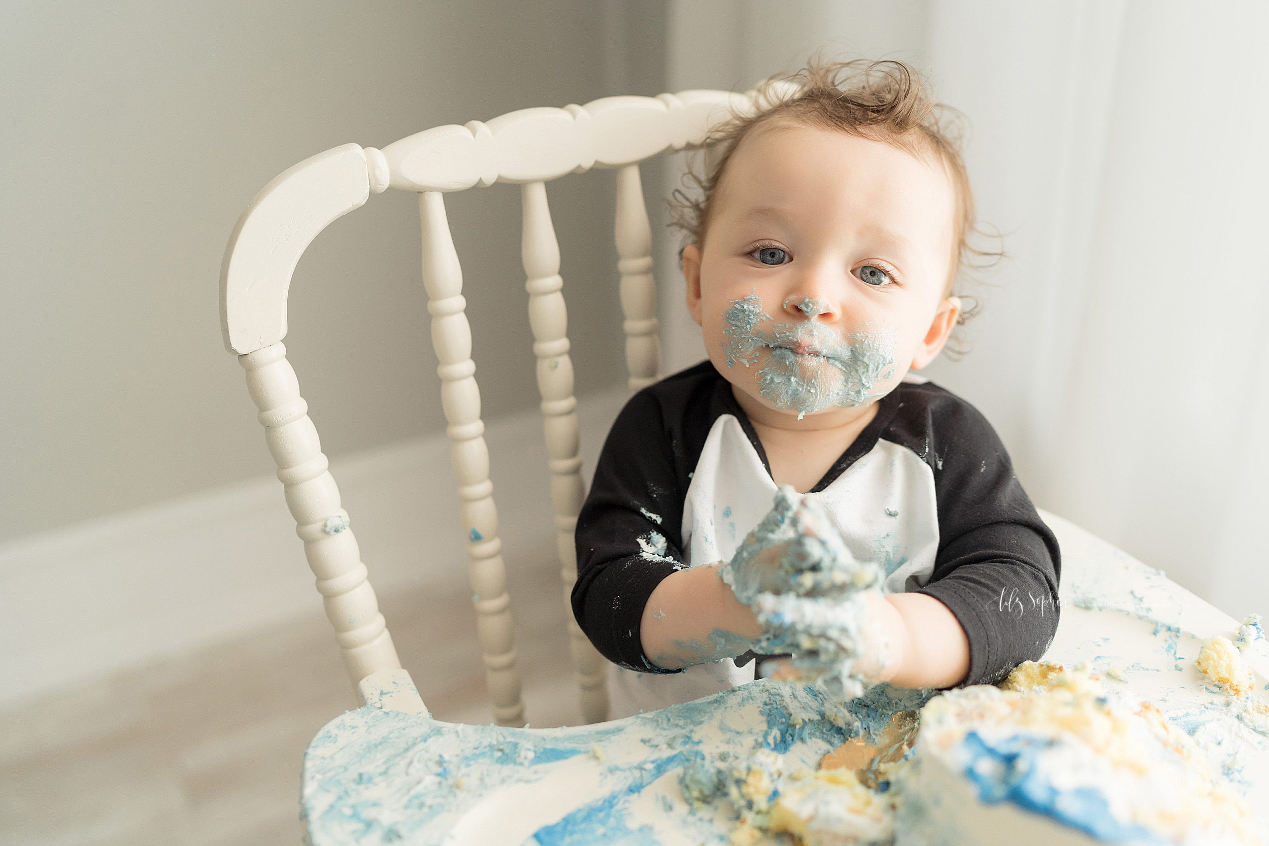  Cake smash photo of a one year old boy taken in a natural light Atlanta studio.  The happy curly haired little boy is sitting in an antique high chair with blue icing on his face and hands and cake and icing smashed all over the high chair tray. 