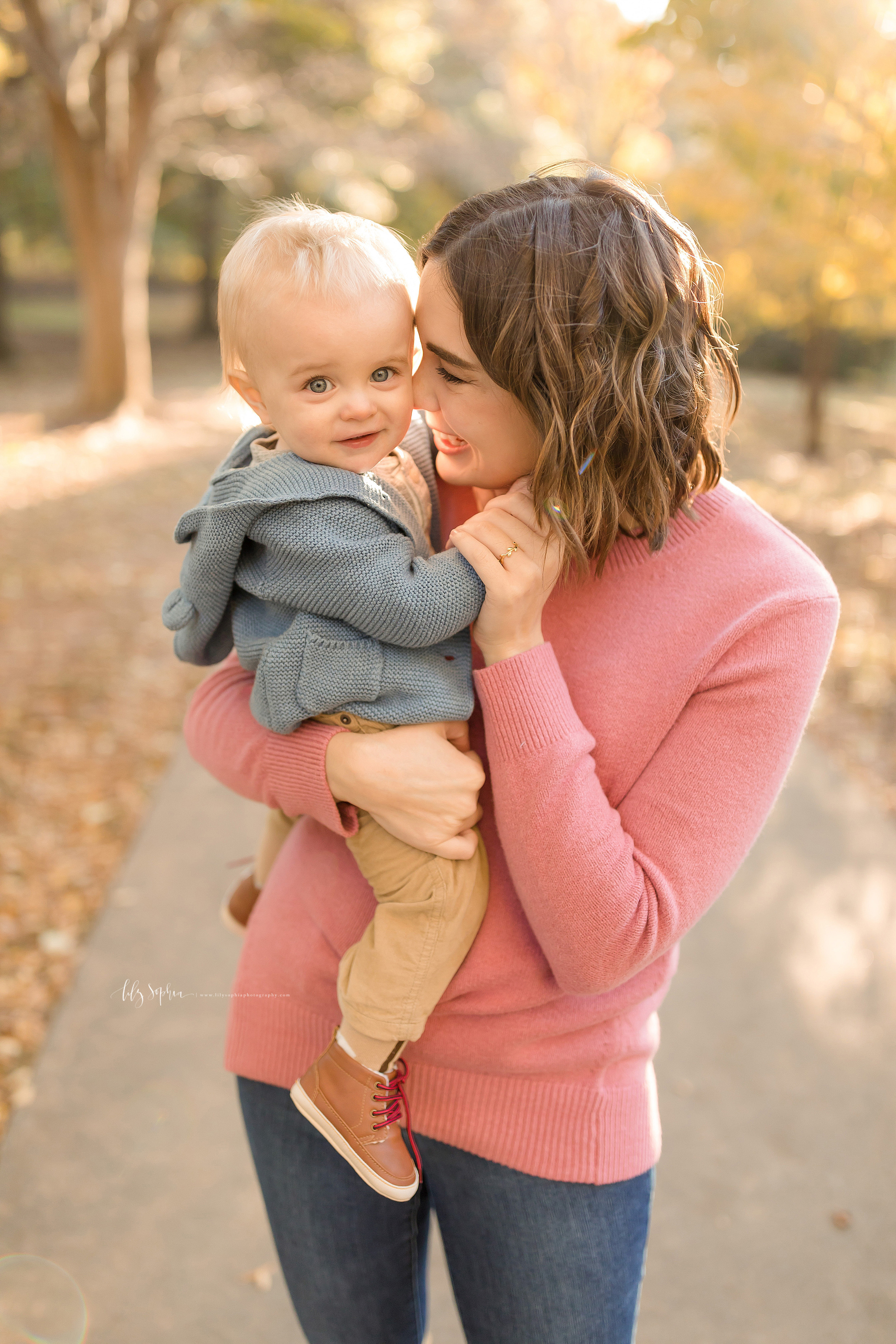  Photo of a happy mom and her son in a park in Atlanta at sunset during the fall taken by Lily Sophia Photography. The brunette Mom is holding her blue-eyed little boy in her arms and is snuggling with him as she stands in a pink long sleeved sweater