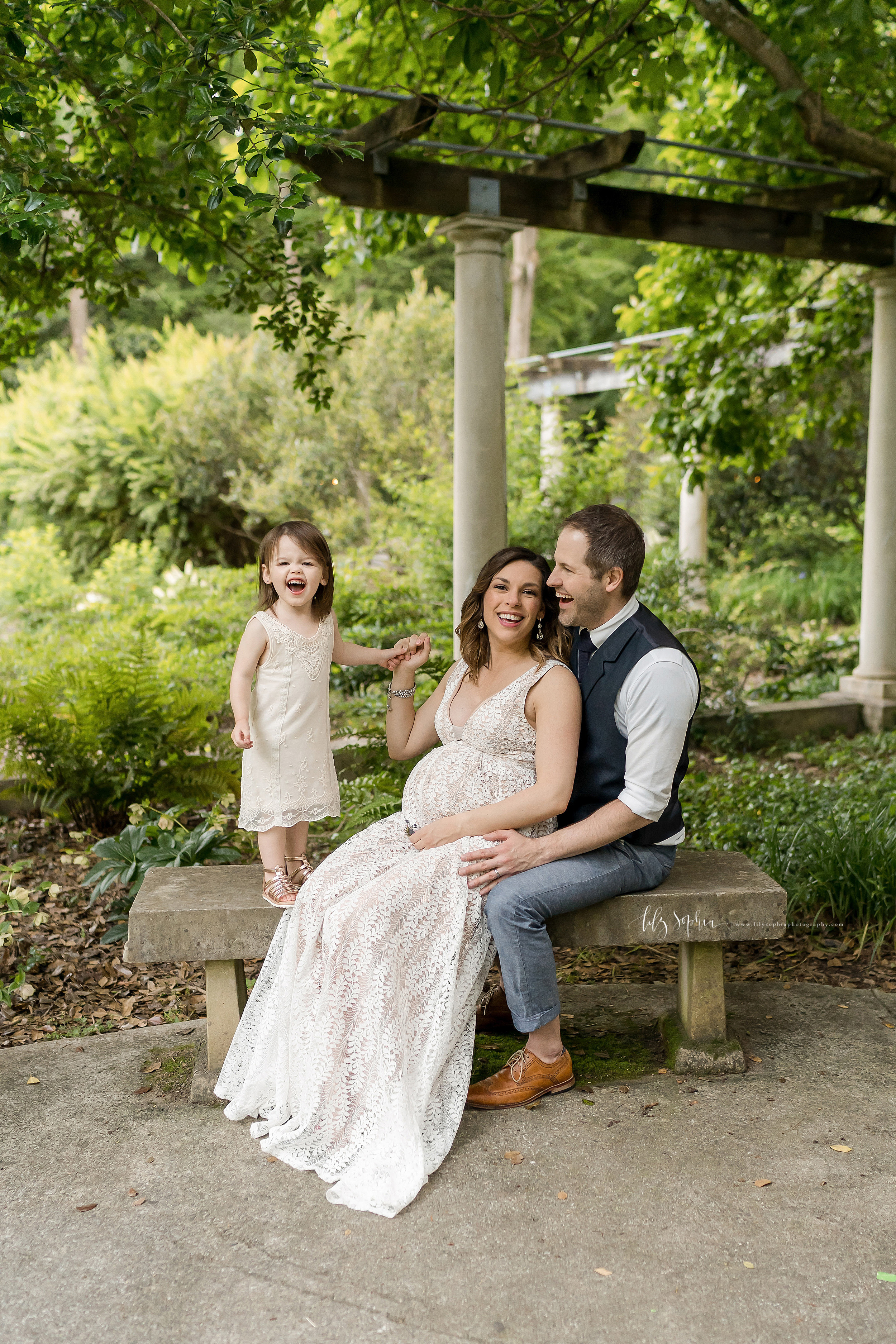  Family photo of a pregnant mom sitting on a stone bench in an Atlanta garden with her back against her husband’s chest and her laughing young daughter stands next to her on top of the bench as she holds her hand. 