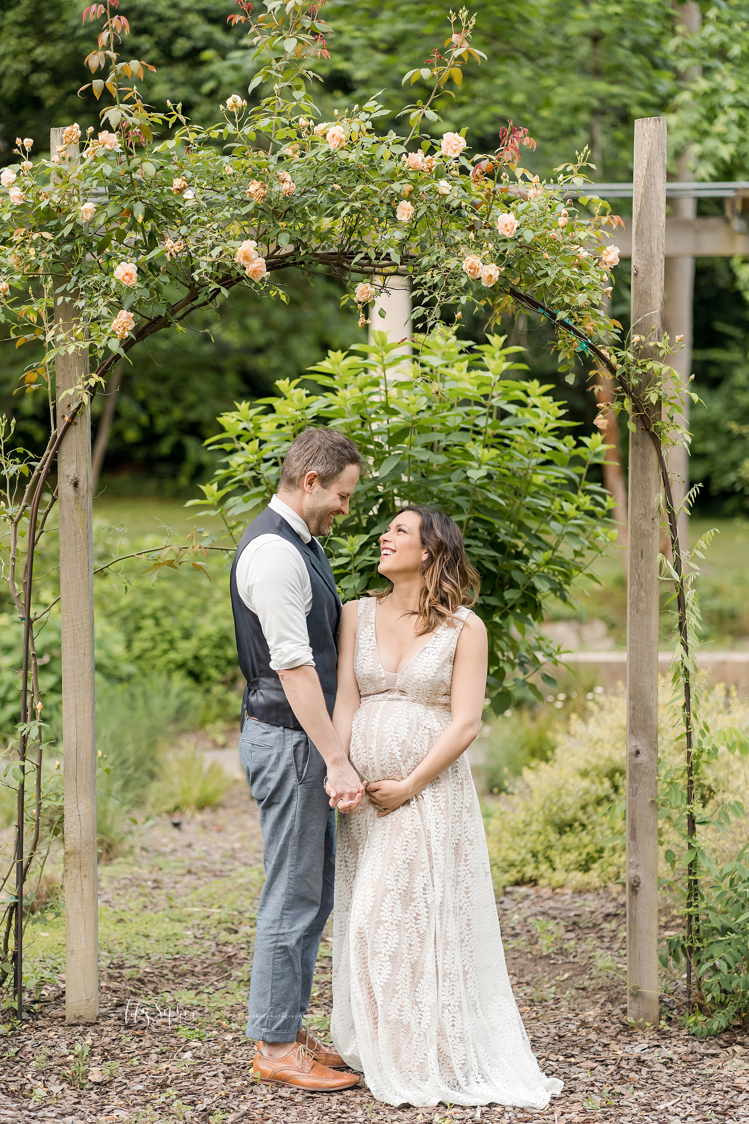  Photo of a pregnant wife with her husband standing and holding hands under an arbor in a garden in Atlanta. 