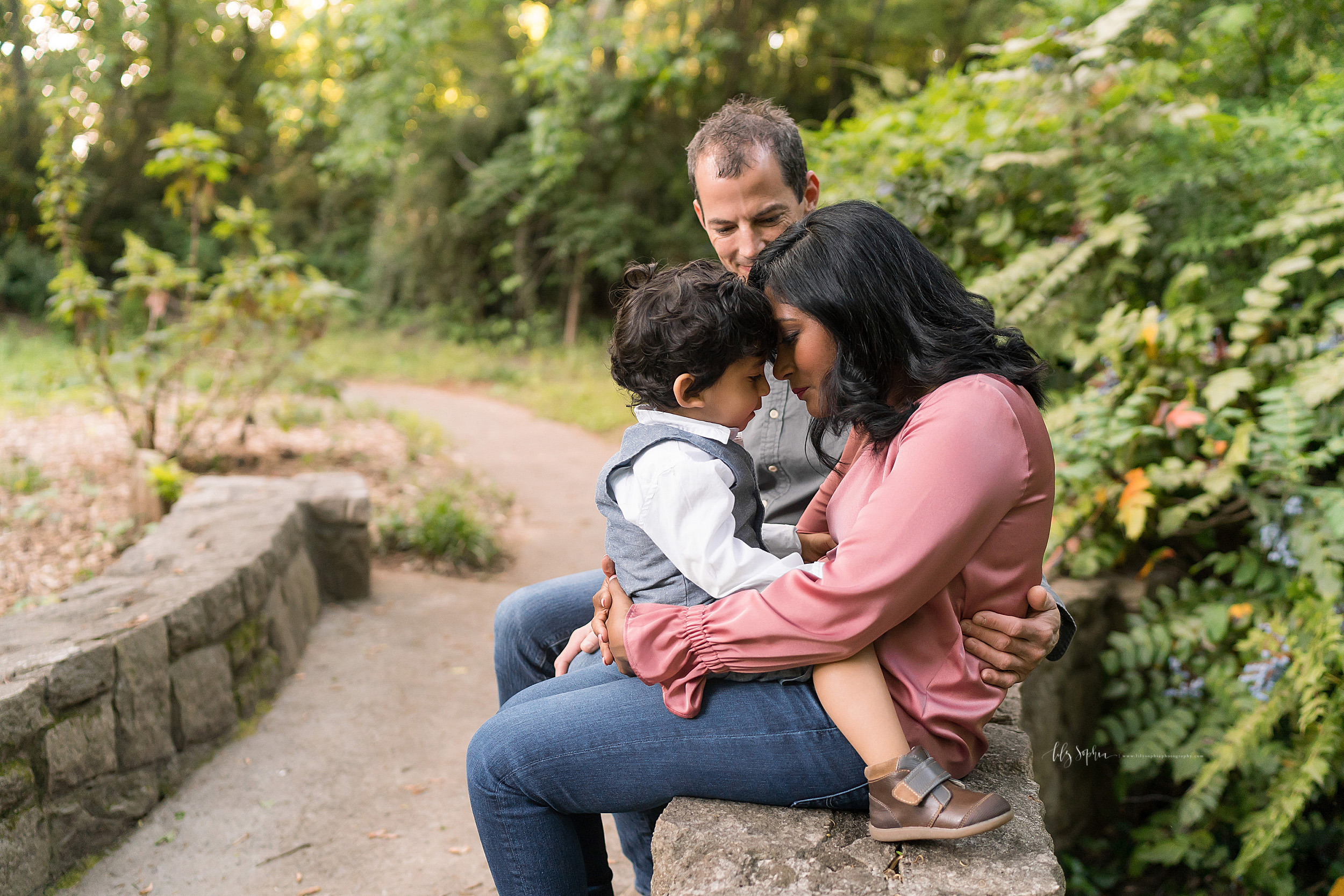  Family photo of three as they sit on a stone bench at sunset in an Atlanta garden with mom and sun putting their heads together and dad looking on. 