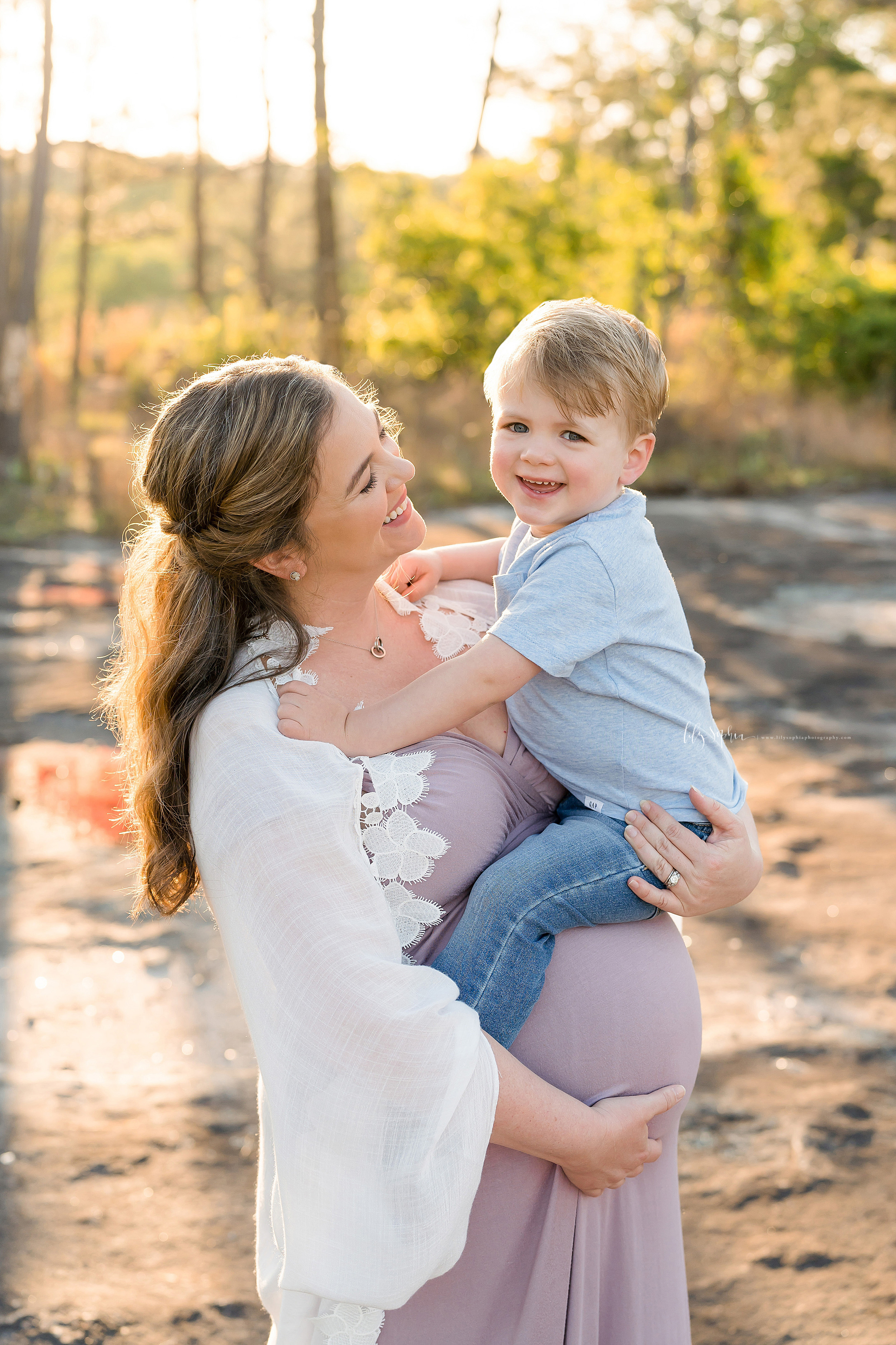  Photo of a smiling mother as she stands holding her toddler son in her right arm and holding her daughter in utero with her left arm on a mountain in Atlanta at sunset. 