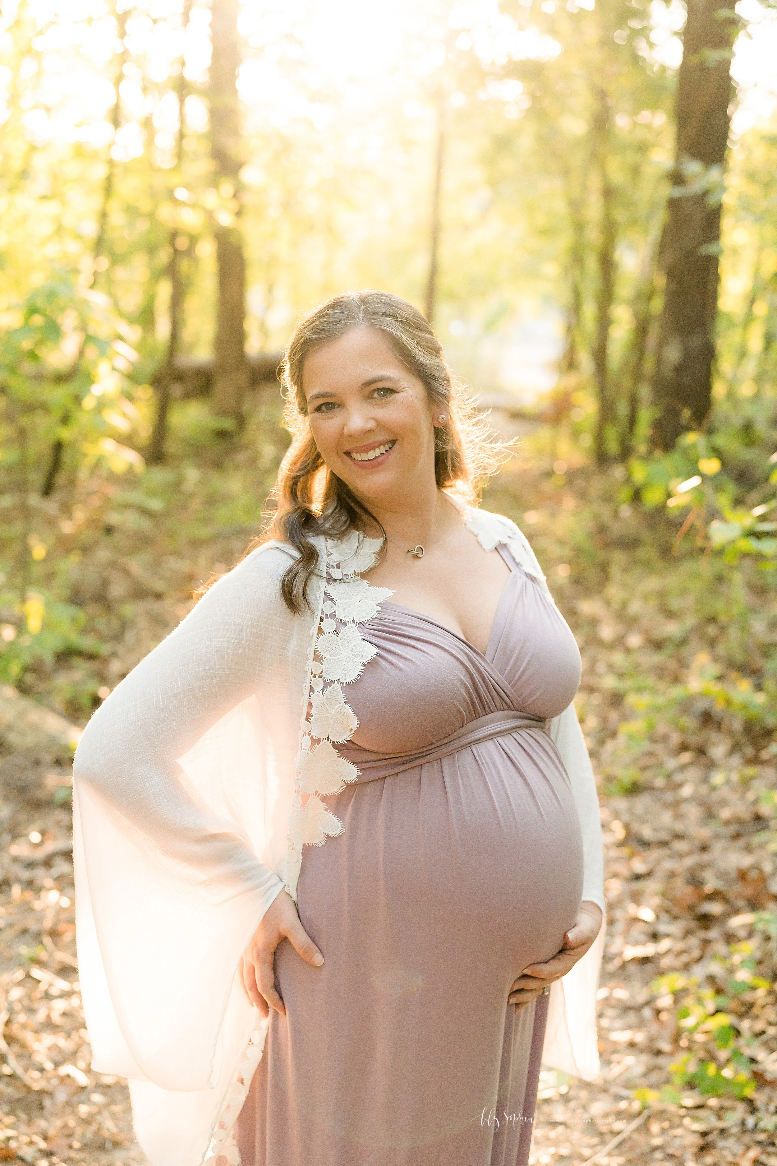  Maternity photo of a blonde woman wearing a purple knit dress with a white shawl as she stands at sunset in the woods on an Atlanta mountain. 