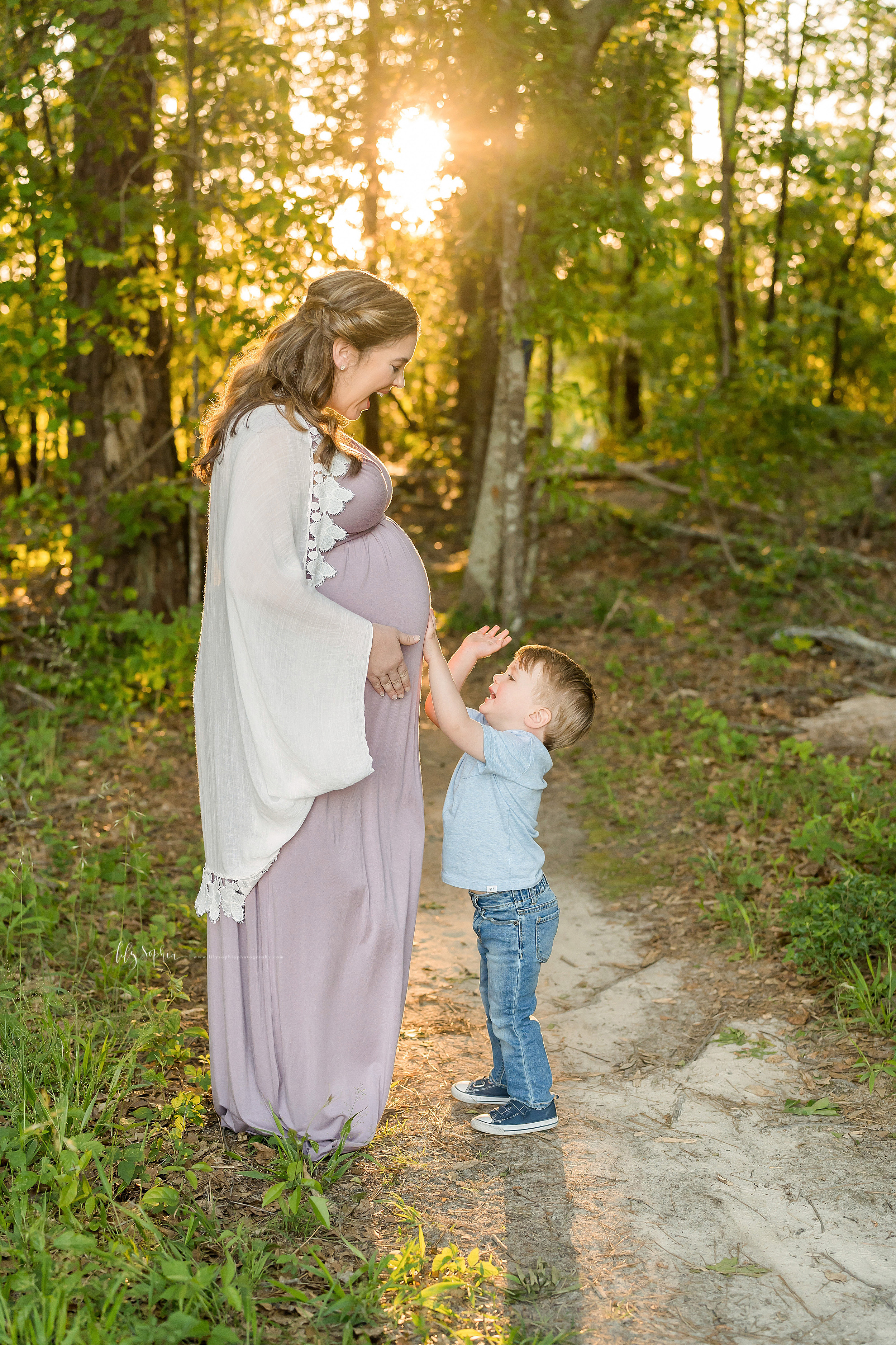  Family photo of a happy mother and toddler son in the woods on a mountain in Atlanta as he plays the drums on her pregnant belly. 