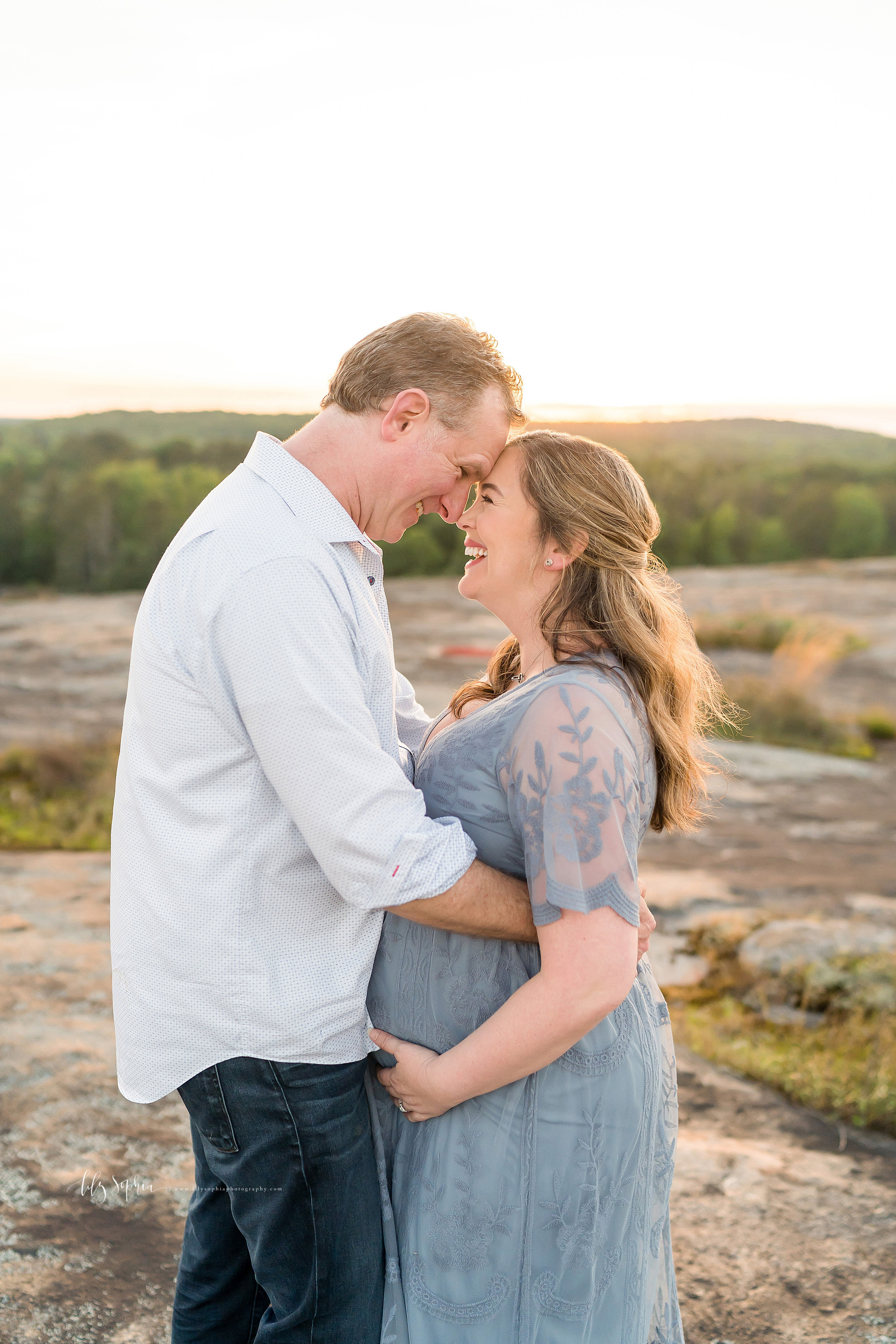 Maternity photo of an ecstatic husband and wife as they laugh with their foreheads together at the top of an Atlanta mountain at sunset.