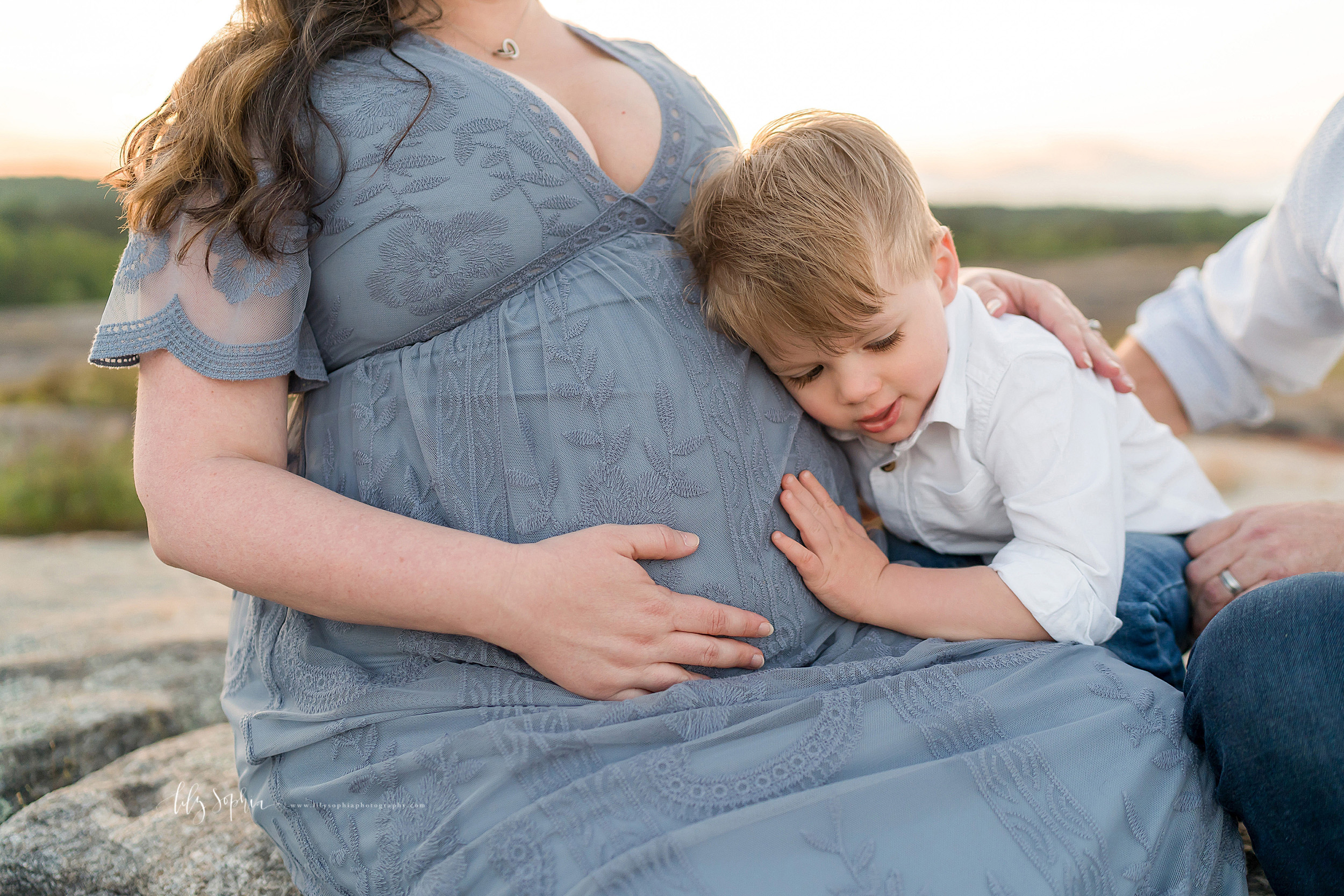  Maternity photo of a family of three on the top of a mountain at sunset in Atlanta, GA.  The toddler boy has his ear to his baby sister in utero as mom and dad sit side by side. 