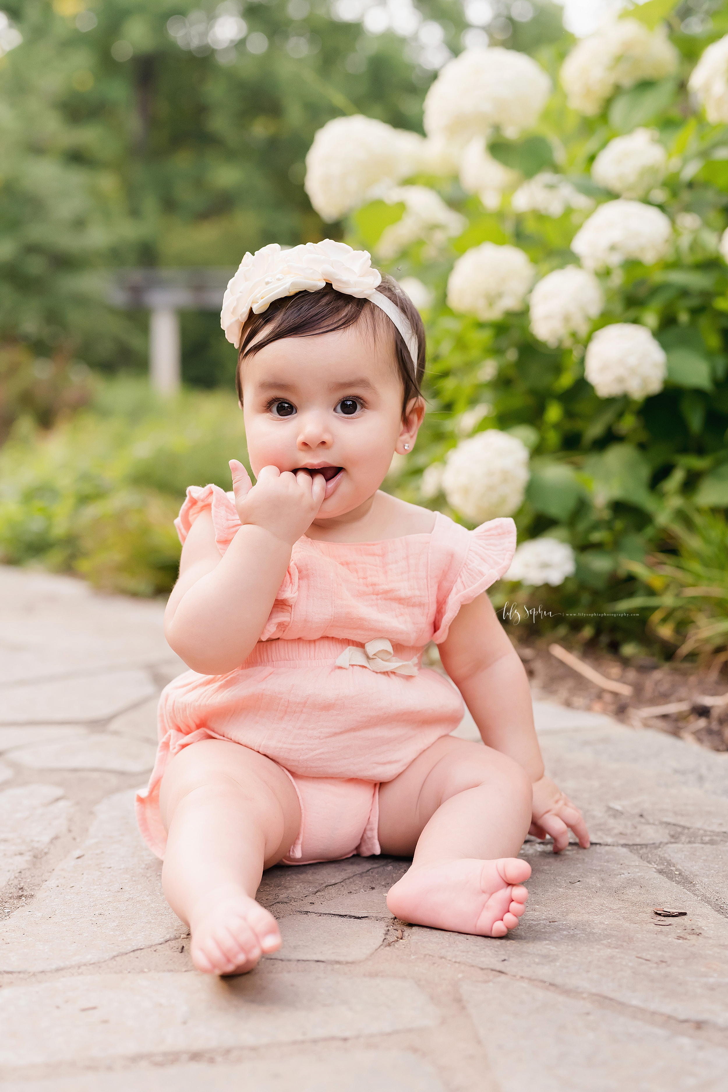  Milestone photo of a baby girl as she sits on a flagstone path and chews on her fingers in an Atlanta garden at sunset. 