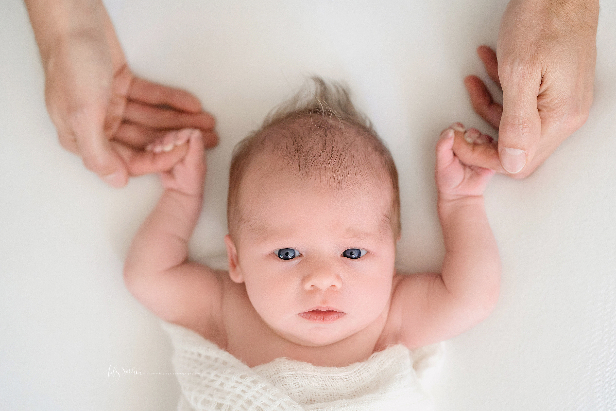  Newborn photo of an alert and wide awake baby girl taken in Atlanta in natural light as her father holds her hands and lifts them over his baby girl’s head. 