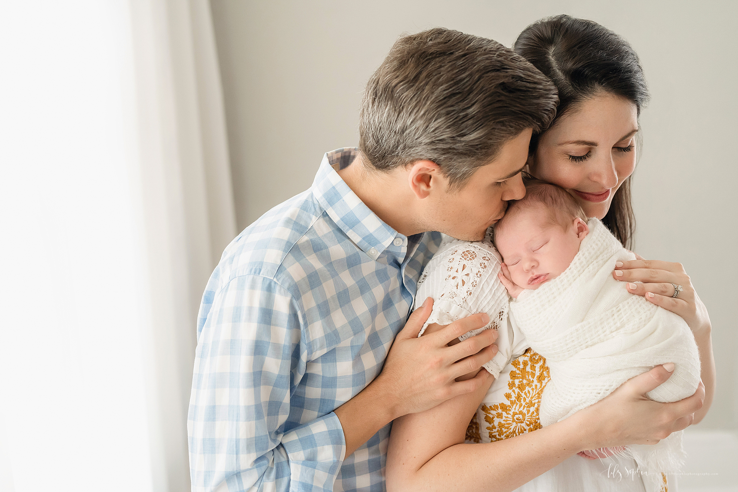  Family photo of a mom and dad snuggling with their contented newborn daughter as they stand in an Atlanta studio holding her. 