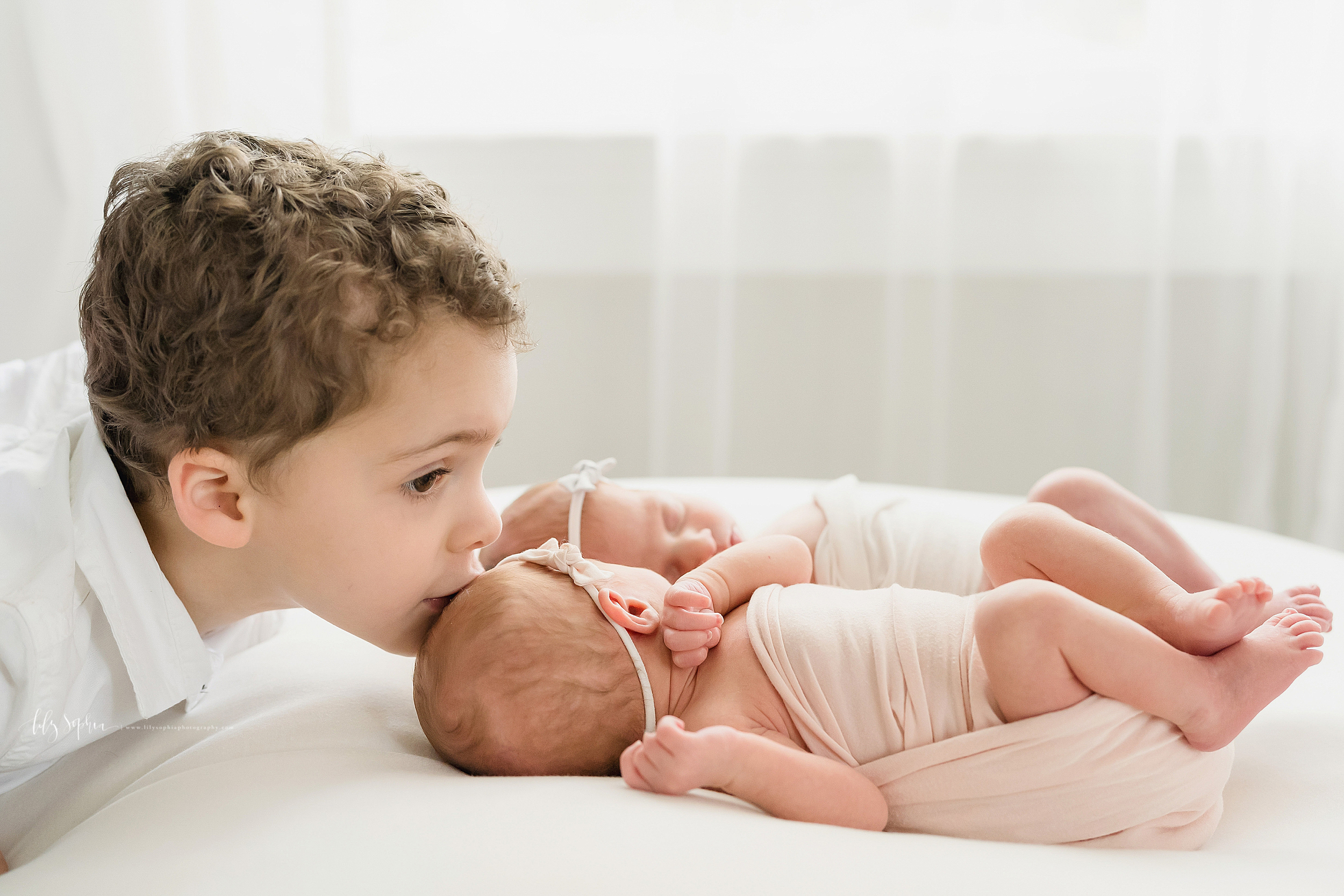  Family photo of a brother and his newborn twin siblings as he kisses the head of his sister while the twins lay on a bed in an Atlanta studio in natural light. 