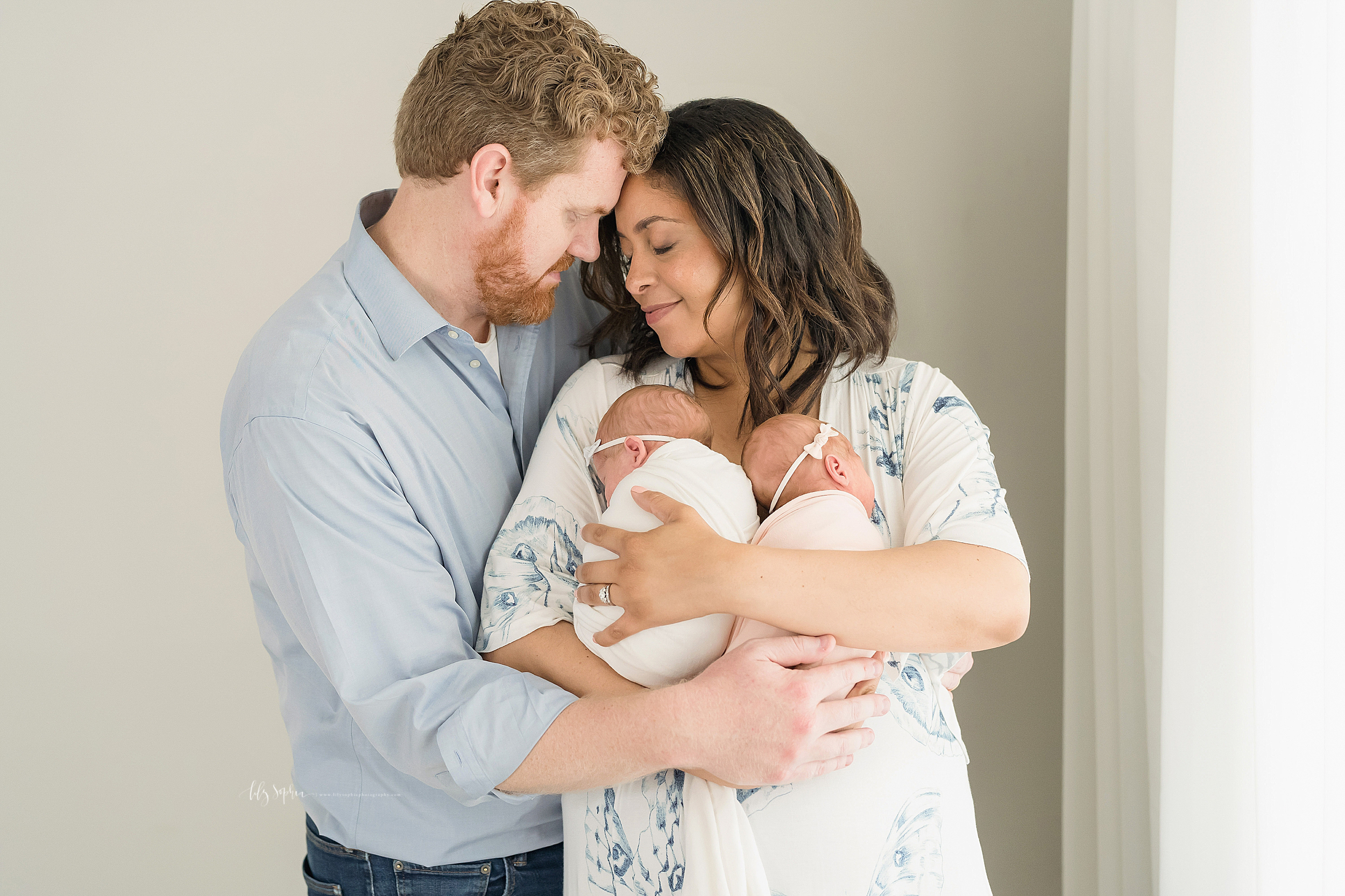  Family photo of a mother and father embracing one another with thankful looks on their faces as mom holds her newborn twins in her arms in a natural light Atlanta studio. 