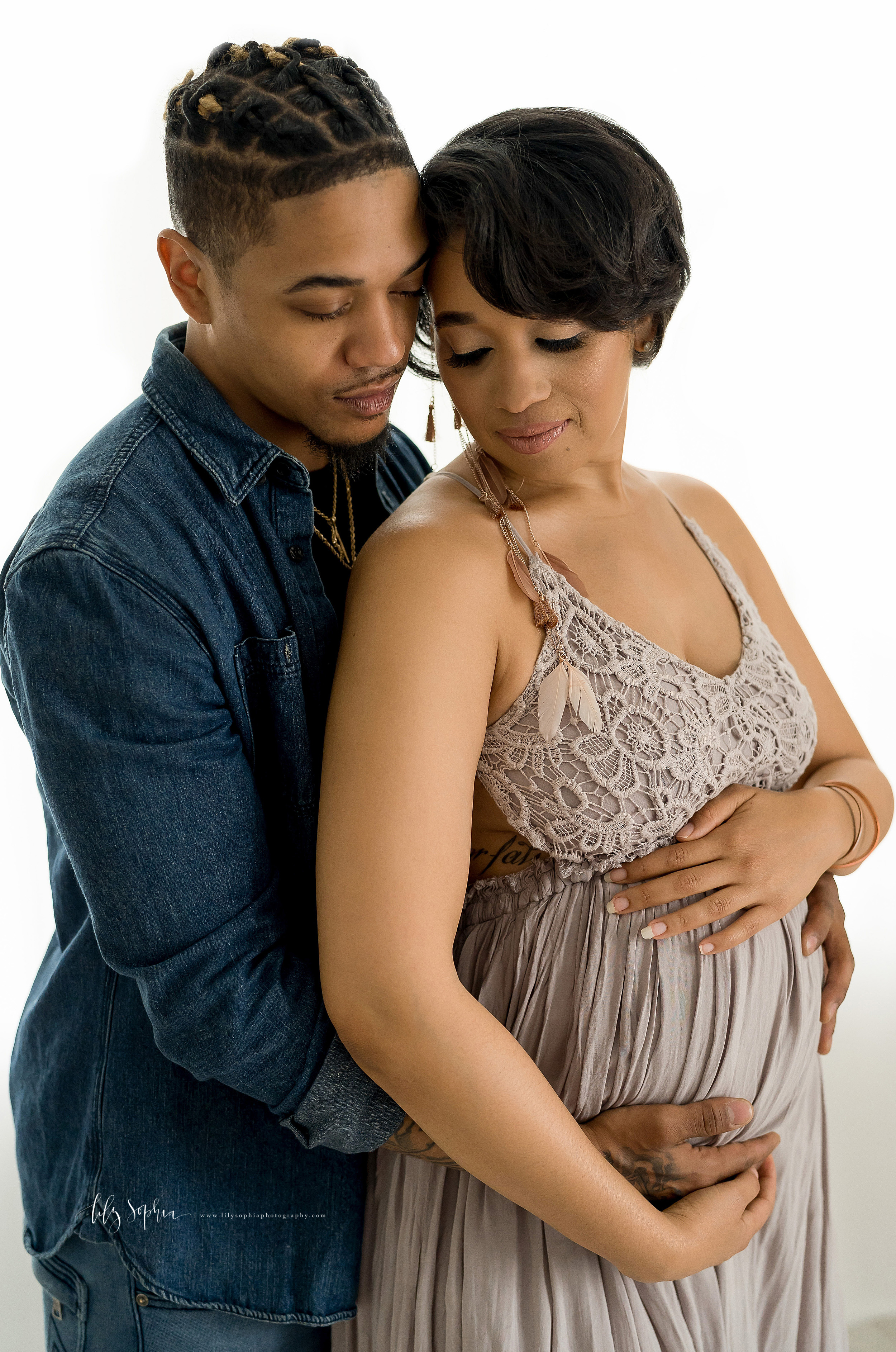  Maternity shoot of an African-American couple in an Atlanta studio with the woman standing in front of her husband as he embraces her from behind and the two of them embrace their child in utero in natural light. 