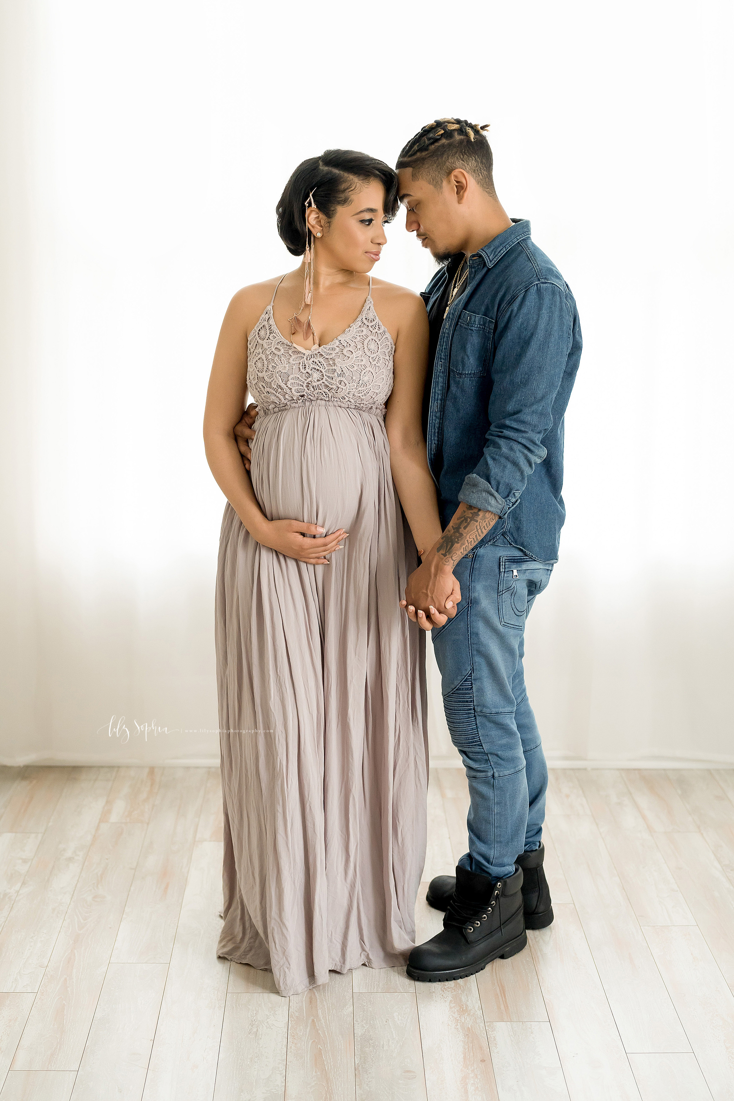  Maternity photo of an African-American woman wearing a lace bodice halter gown as she stands with her left shoulder to her husband’s chest, holds her baby in utero with her right hand, and holds her husband’s hand with her left hand as they put thei