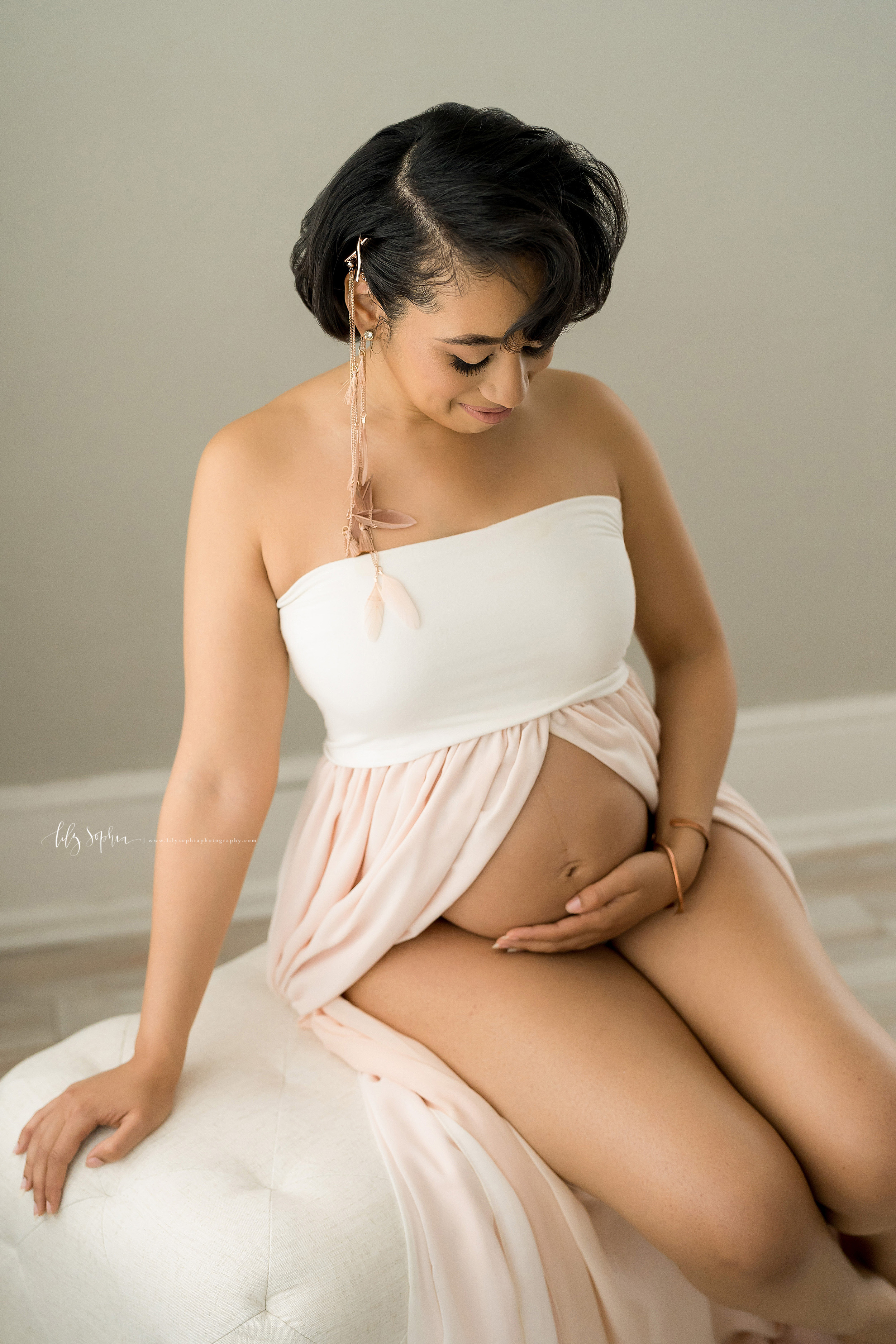  Pregnant African-American woman sits on a bed in a sleeveless bandeau split-front gown and bares her belly to admire her newborn in utero in a natural light Atlanta studio for her maternity shoot. 