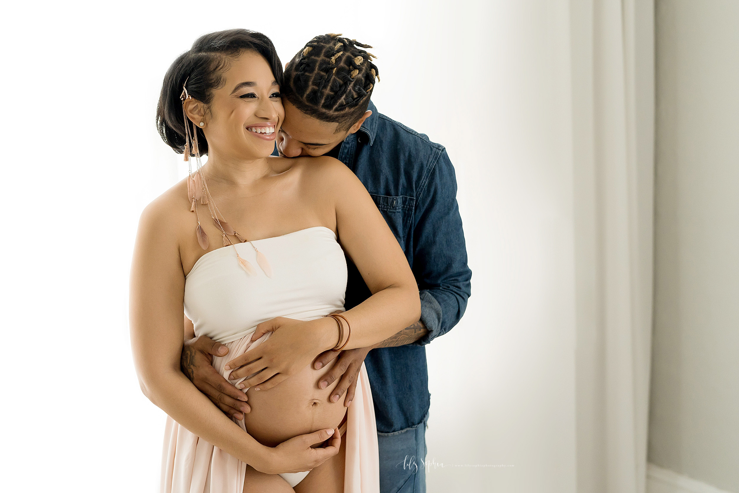  Smiling pregnant African-American woman stands in a bandeau split-front gown as her husband kisses her on her shoulder and the two of them embrace her bare pregnant belly in an Atlanta studio using natural light photography. 