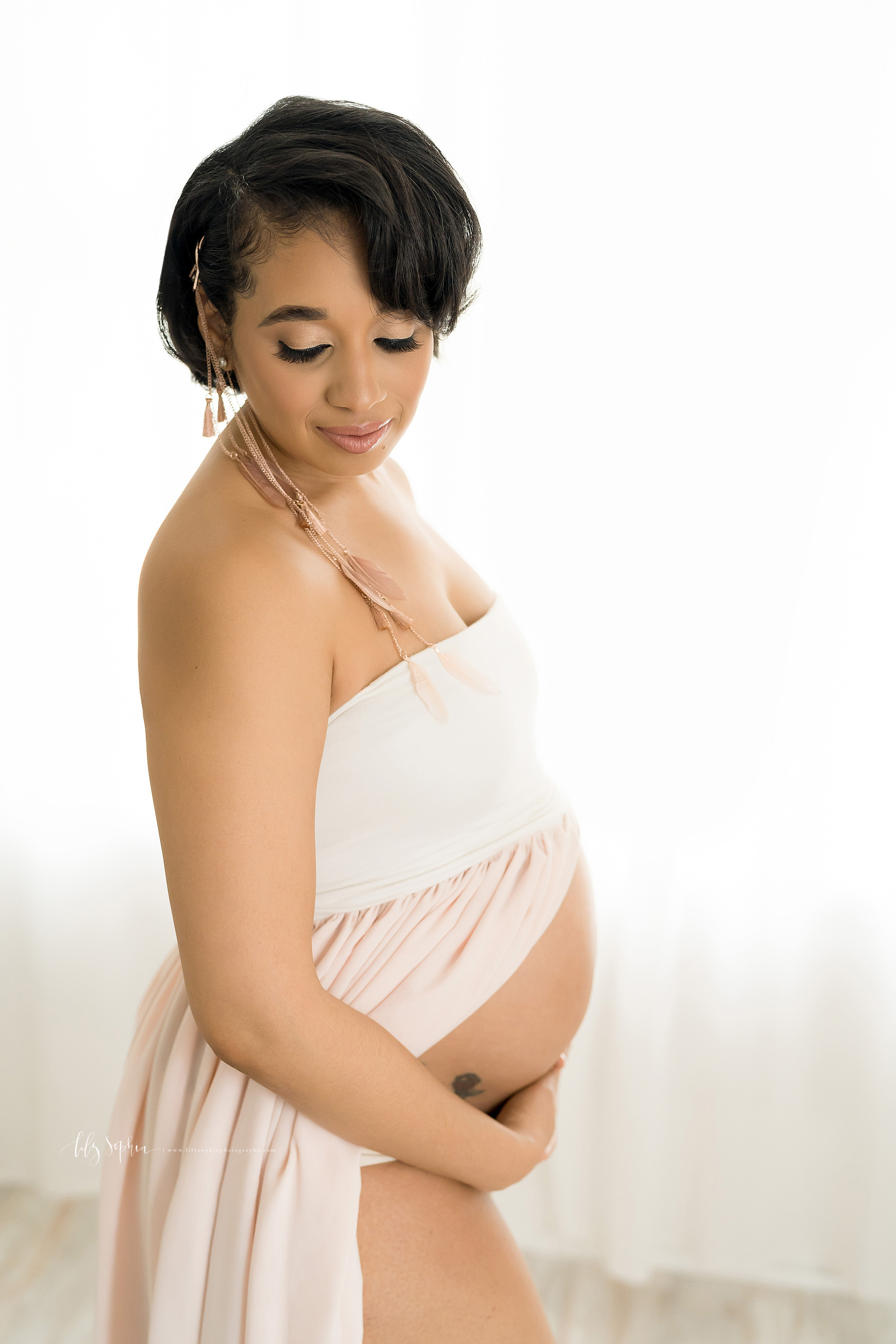  Maternity photo in an Atlanta natural light studio of a contemplative African-American mother as she stands wearing a white and rose bandeau split front gown and holds her bare belly.  