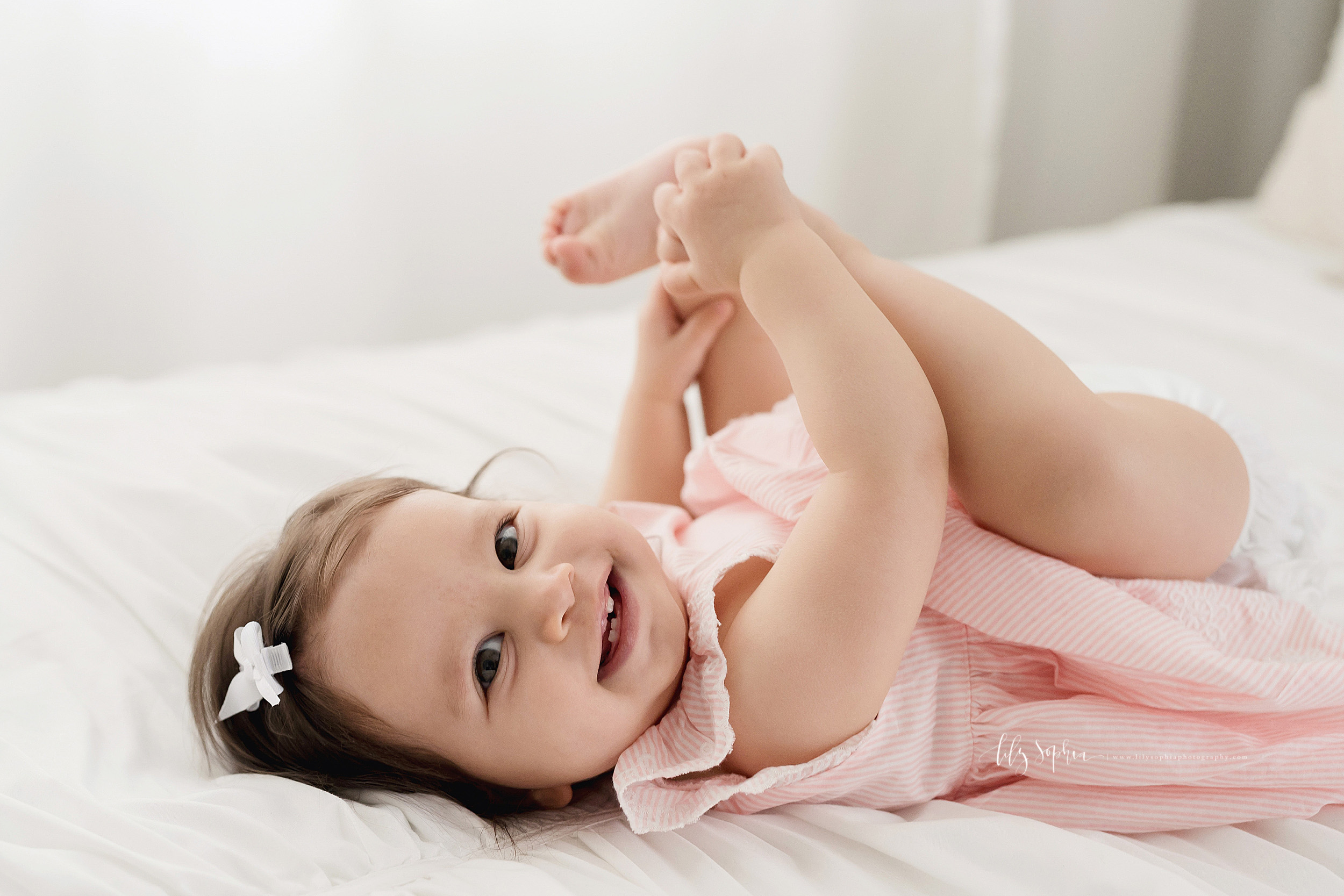  Milestone photo of a smiling nine month old baby girl as she lies on her back on a bed in a natural light Atlanta studio and plays with her toes. 