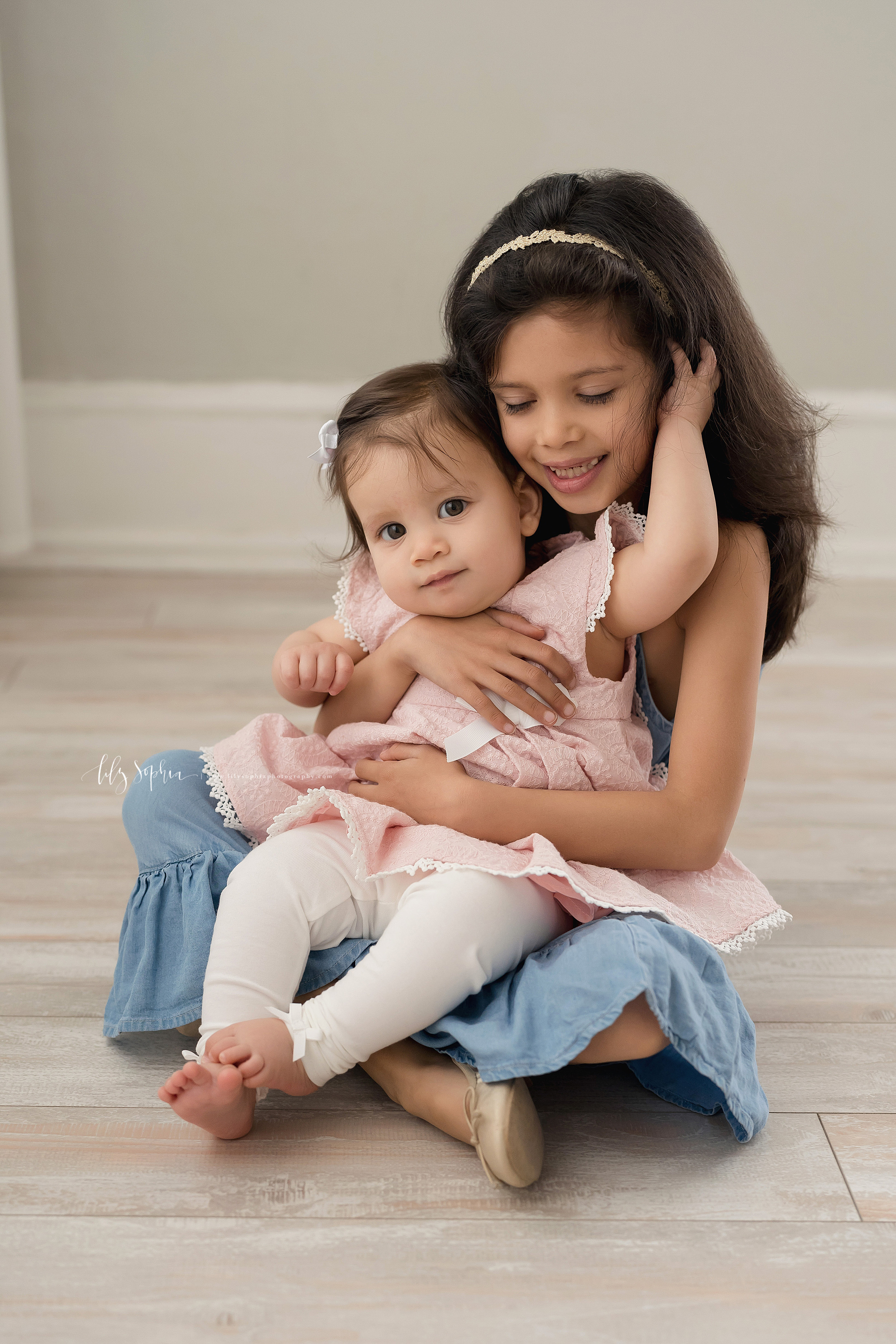  Sibling photo of two sisters as the older sister sits with her nine month old baby sister on her lap adoring her as her baby sister plays in her hair and they sit on the floor of an Atlanta studio in natural light. 