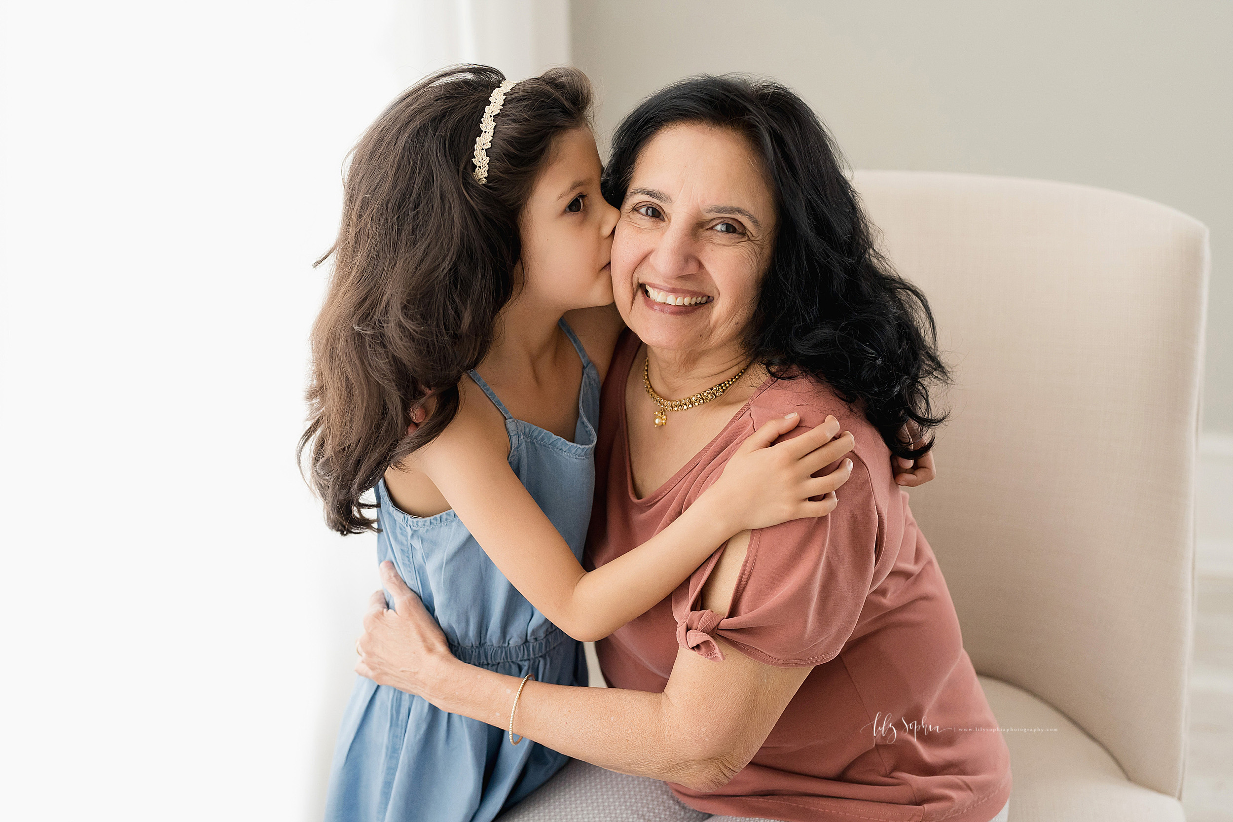  Family photo of a granddaughter standing next to her grandmother in a natural light studio as the two embrace and the granddaughter kisses her grandmother on the cheek in Atlanta, Georgia. 