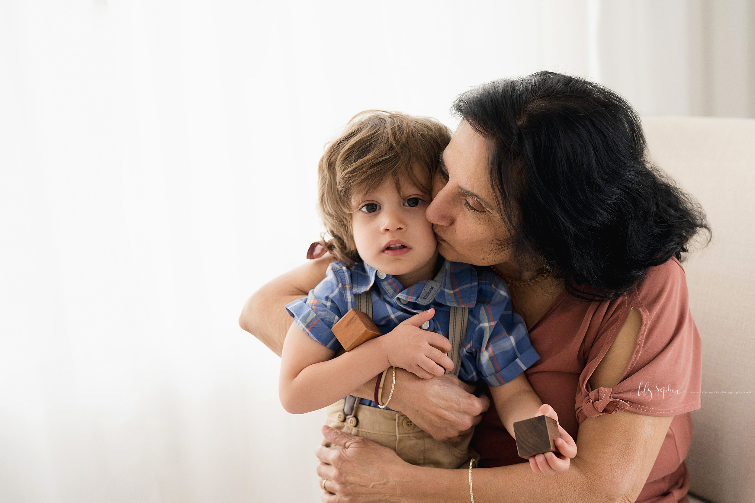  Family photo of a grandmother holding her grandson on her lap in an Atlanta studio as she hugs and kisses her grandson in natural light and he plays with wooden blocks. 