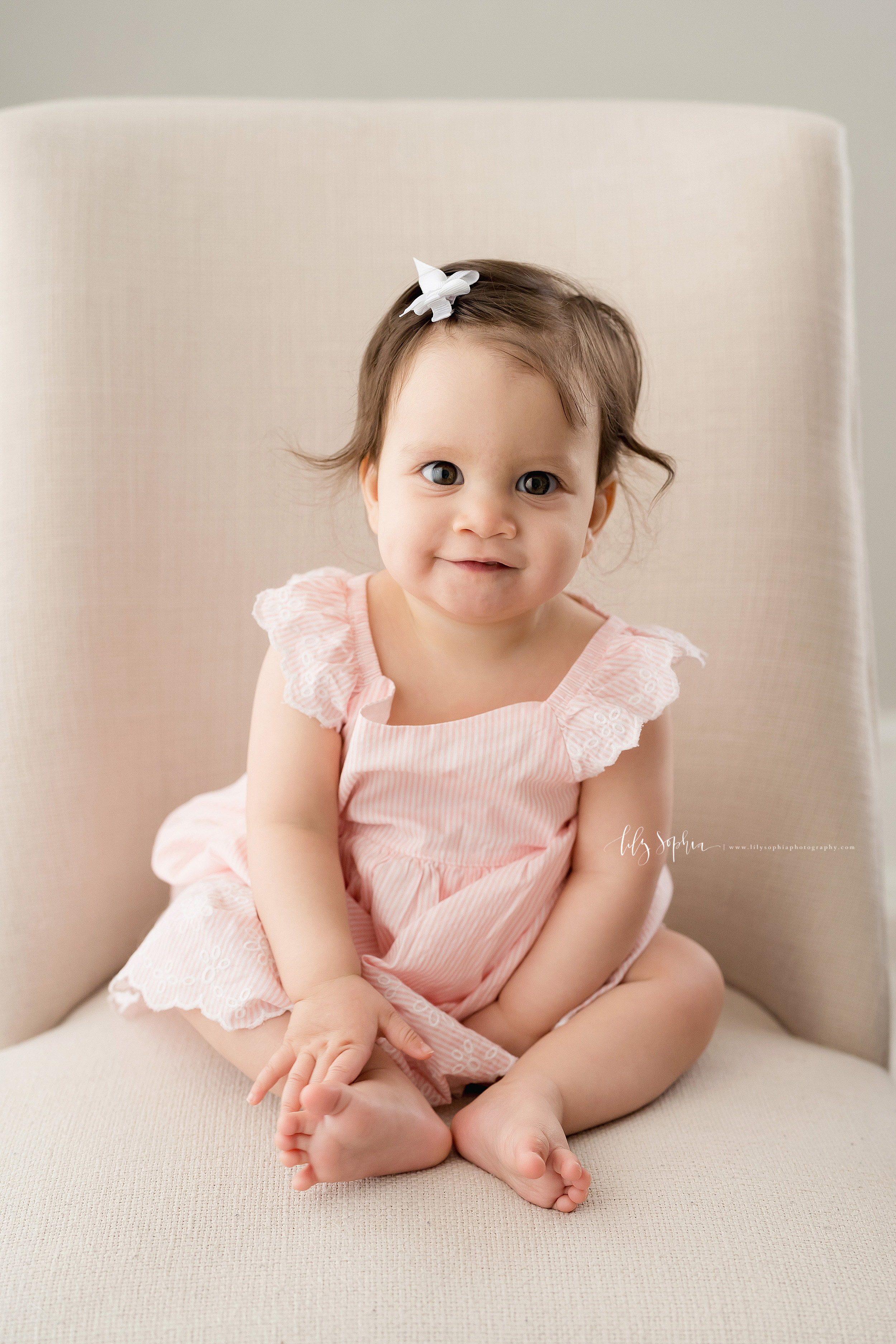  Milestone photo of a nine month old baby girl as she sits in a tan armless chair in natural light in an Atlanta studio. 