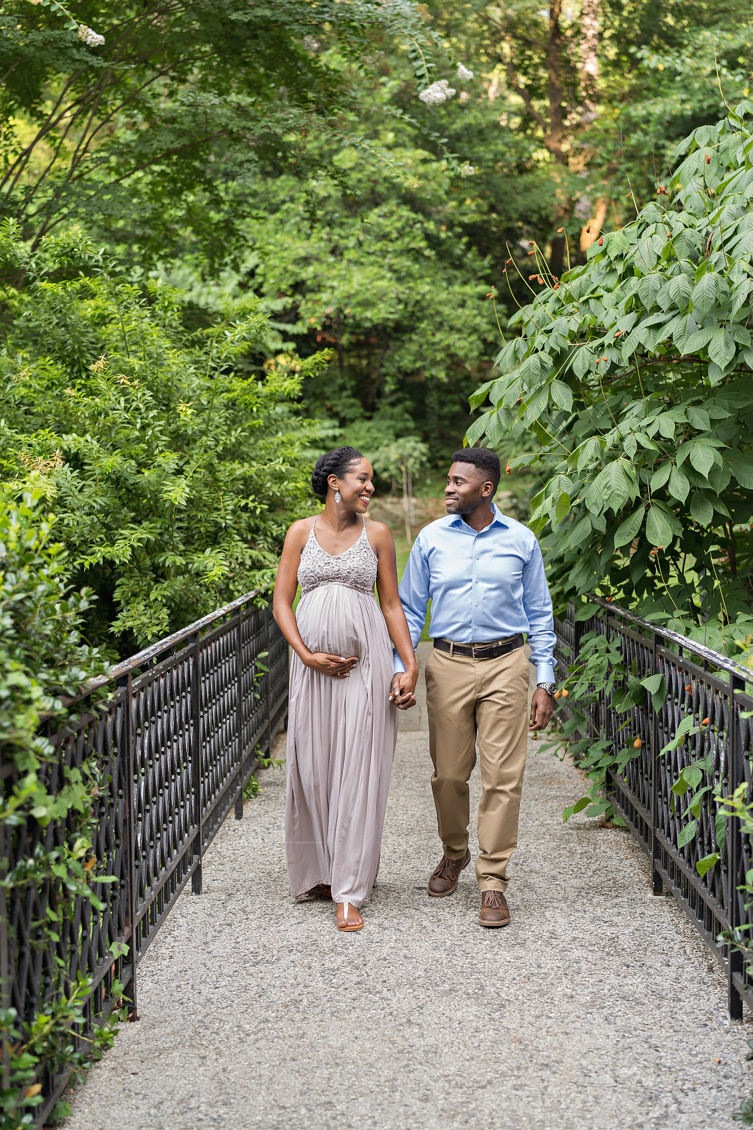  Maternity photo of an African-American husband and wife as they walk hand in hand over a wrought iron bridge at sunset in an Atlanta garden. 