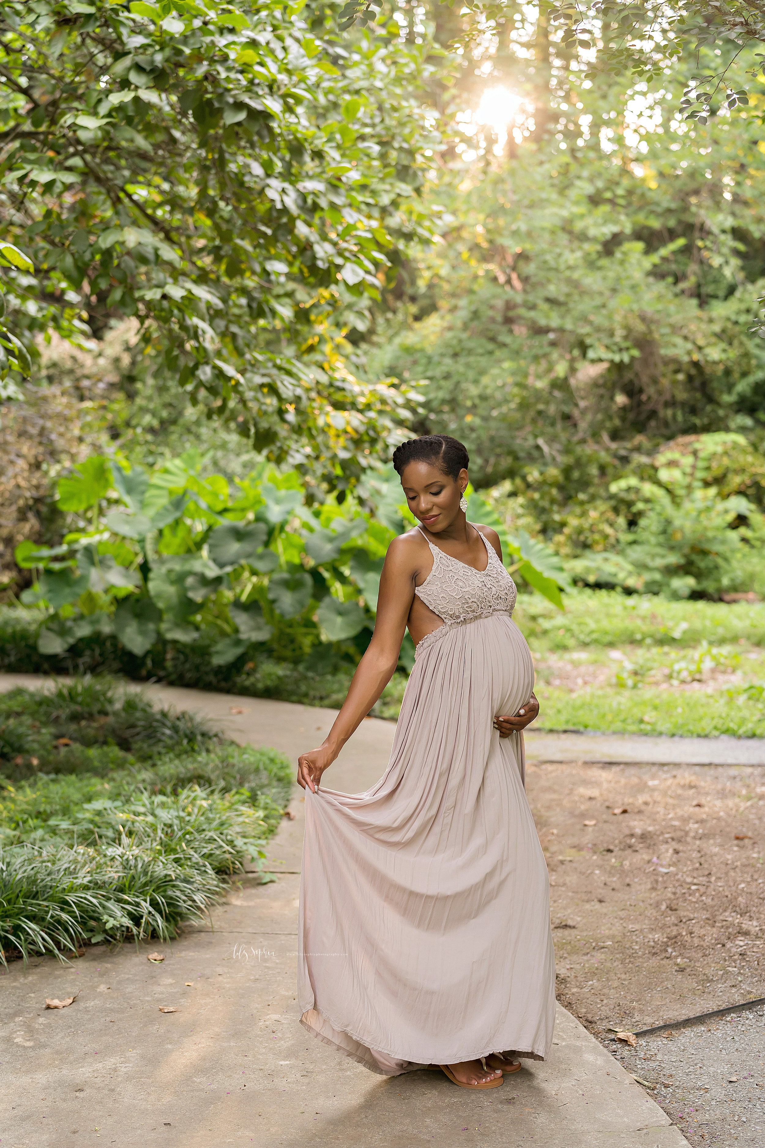  Maternity photo of an African-American mother as she holds her full-length gown and the base of her belly while standing at a cross path in an Atlanta garden at sunset. 