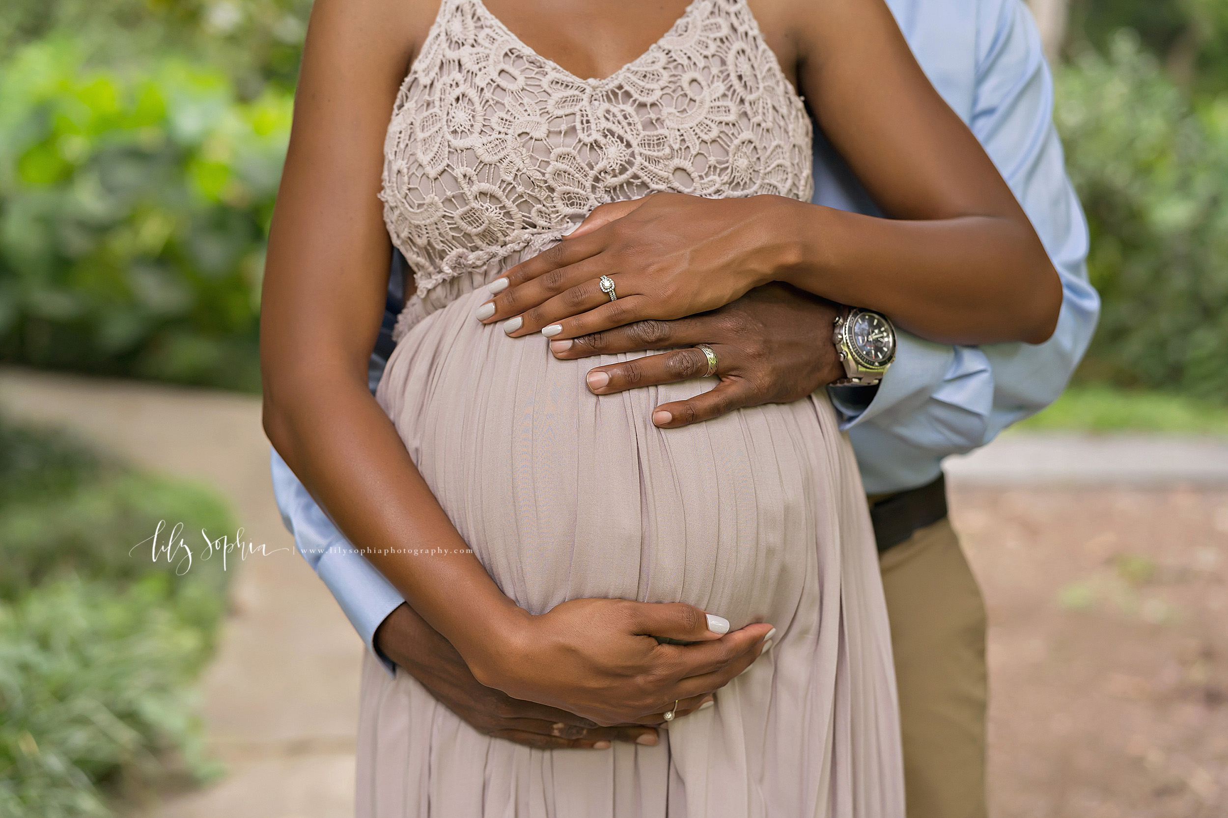  Maternity photo of a close-up of the belly of an African-American woman as her husband stands behind her in an Atlanta garden and the two of them surround their newborn in utero with their hands to demonstrate their love. 