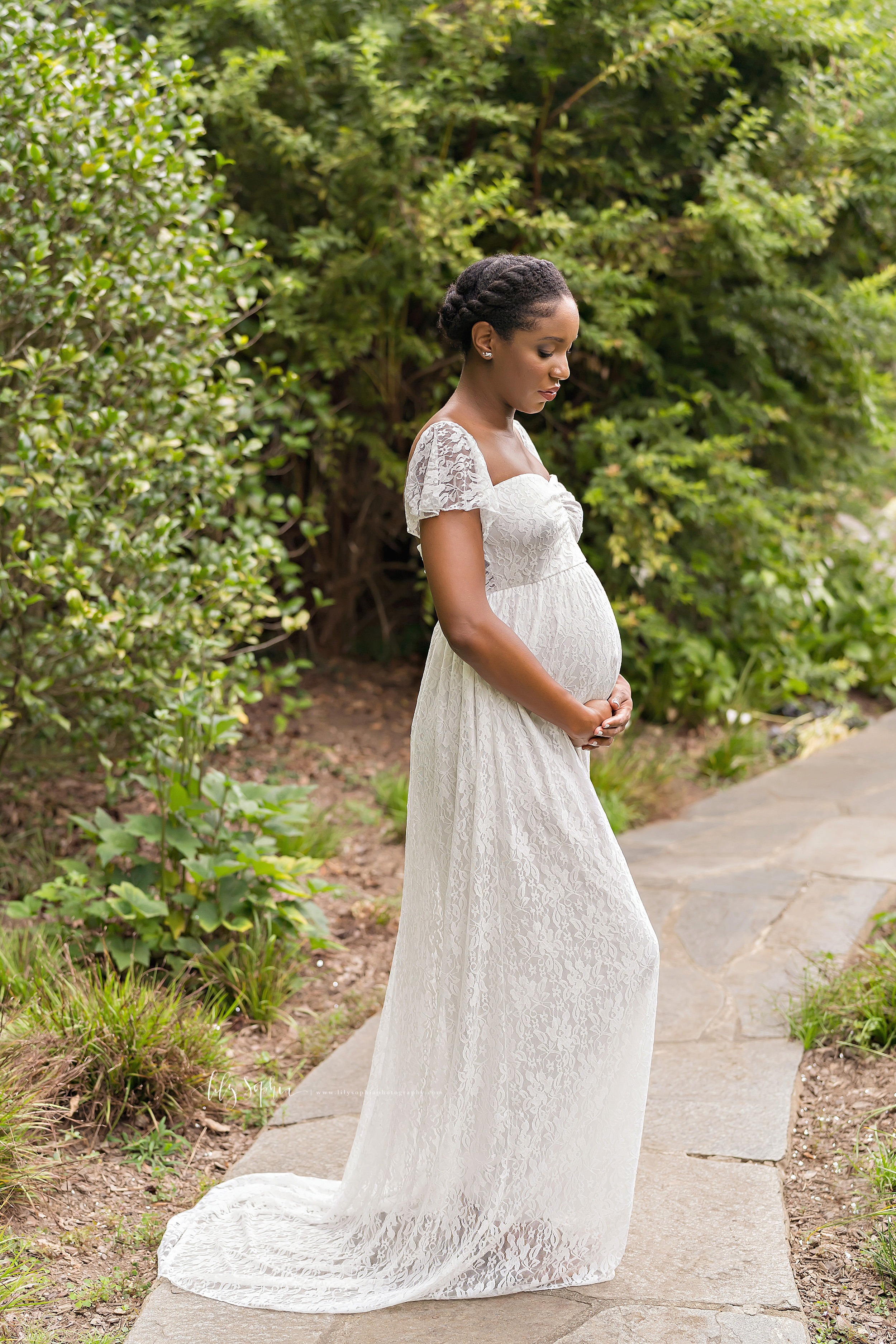  Maternity photo of a contemplative African-American mother as she stands on a stone path in an Atlanta garden and embraces her newborn in utero by holding the base of her belly. 