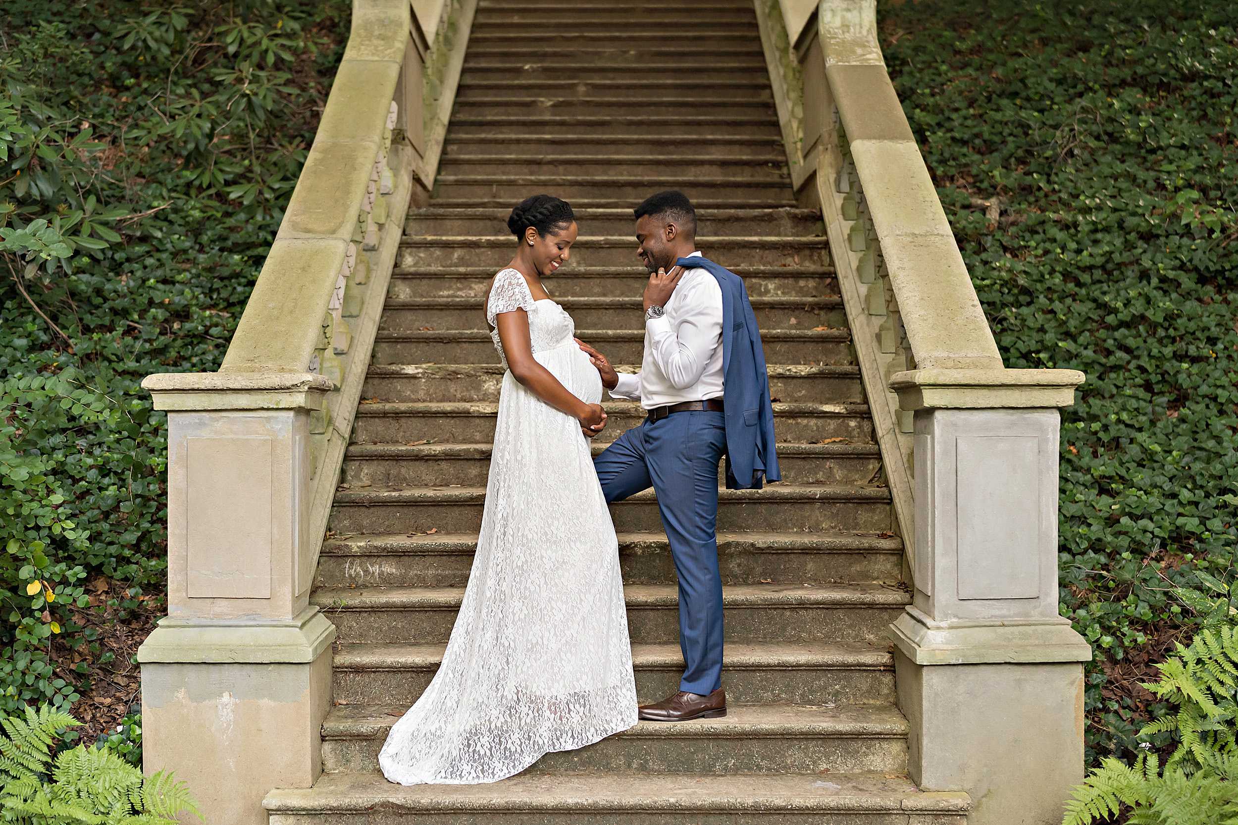  Maternity photo of an African-American husband and wife as they stand at the base of a stone staircase in an Atlanta garden at sunset with the father holding his blue suit jacket over his shoulder and touching his newborn in utero and his wife clasp
