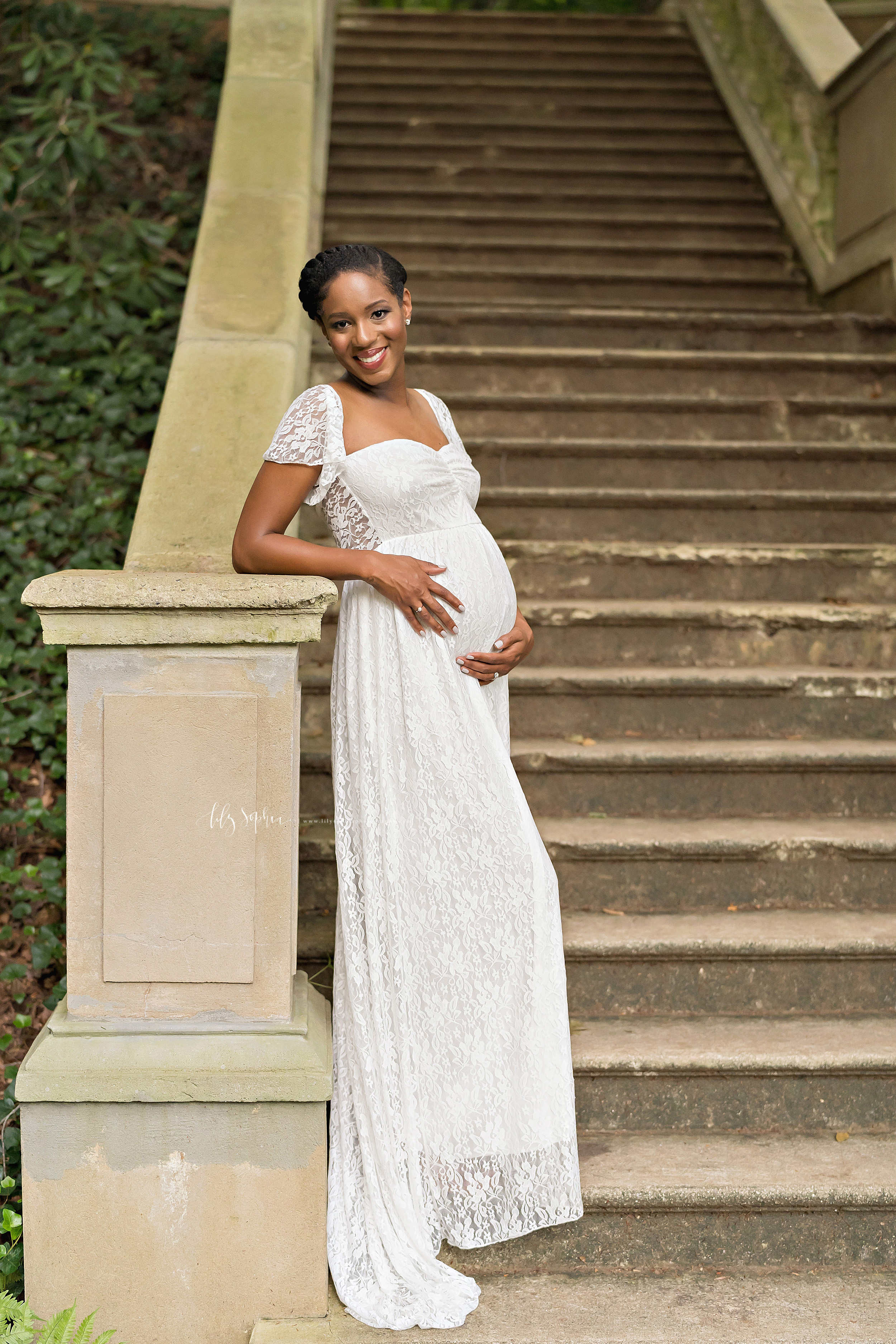  Maternity photo of an African-American woman wearing a lace overlay full-length gown with short bell sleeves as she stands at sunset at the base of a stone staircase and leans with her right elbow on the newel post of the stone bannister in an Atlan