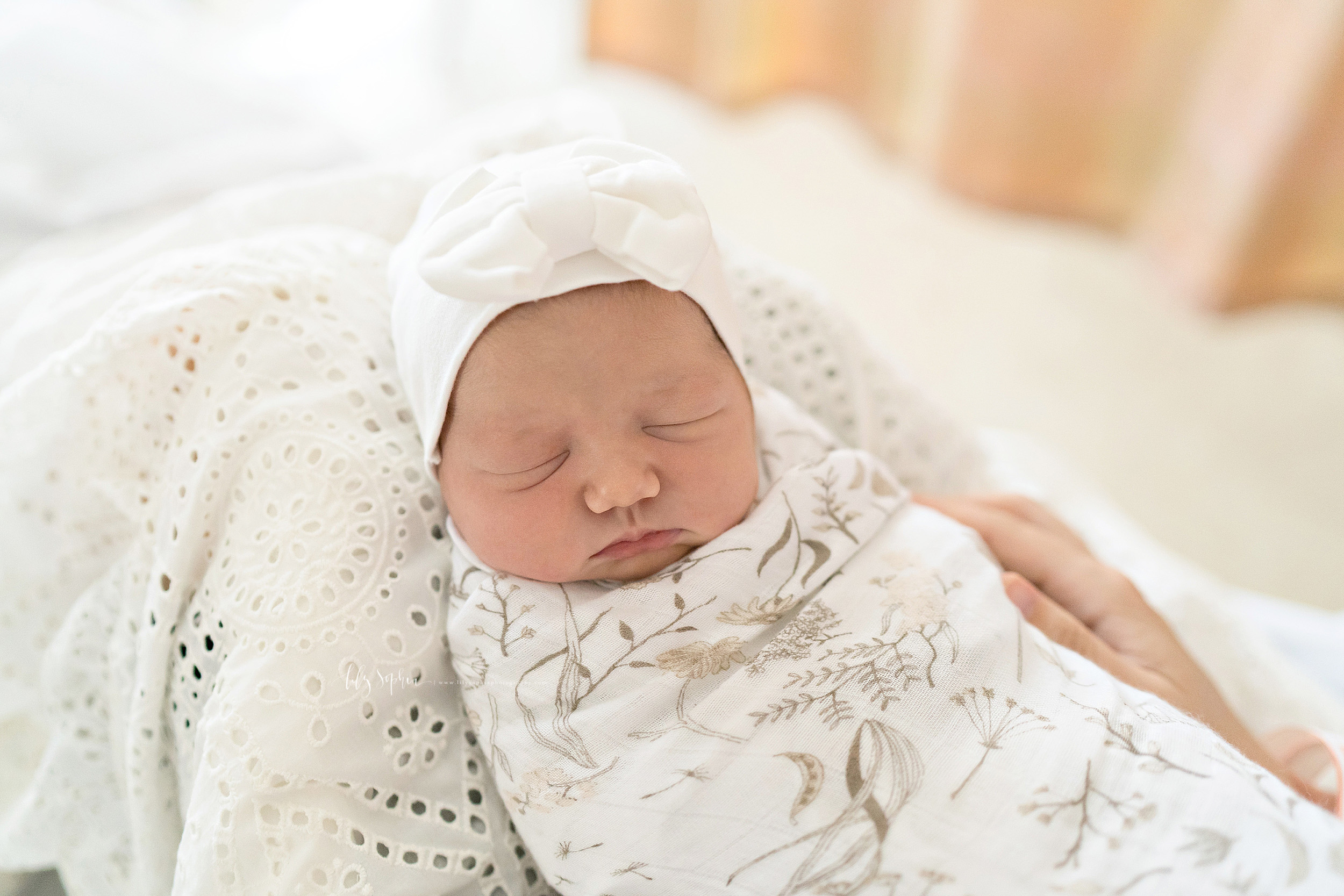  Photo of a sleeping newborn baby girl swaddled to her neck and wearing a cap with a big white bow as she lays against the knees of her mother taken by Lily Sophia Photography in Manhattan, New York. 